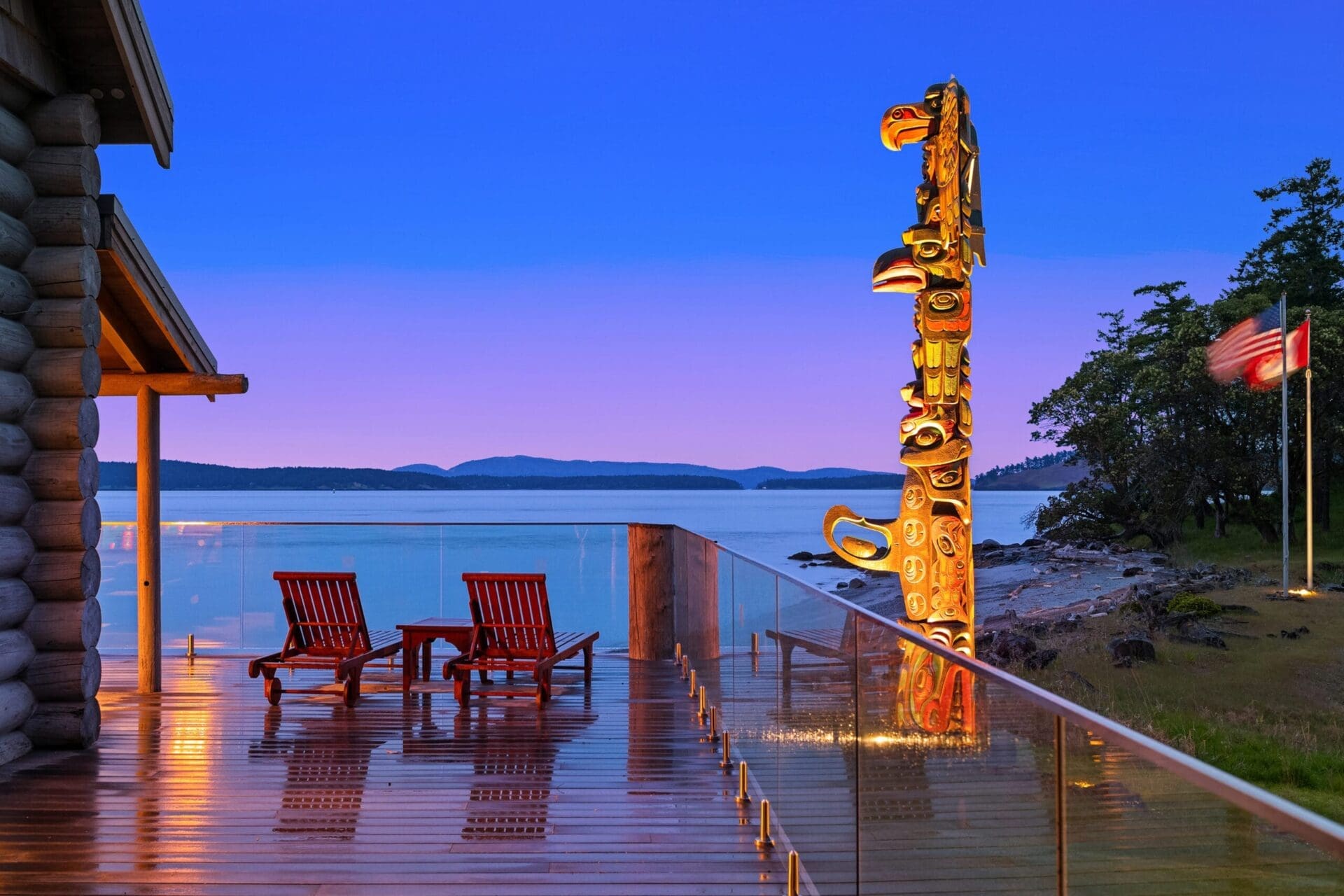 Twilight replacement gallery2 A serene lakeside scene at sunset features an hdr wooden deck with two empty lounge chairs, a totem pole, and a reflecting glass railing. The calm lake and distant mountains enhance the peaceful atmosphere, with a wooden cabin partially visible on the left. Perfect for top real estate listings. Twilight replacement.