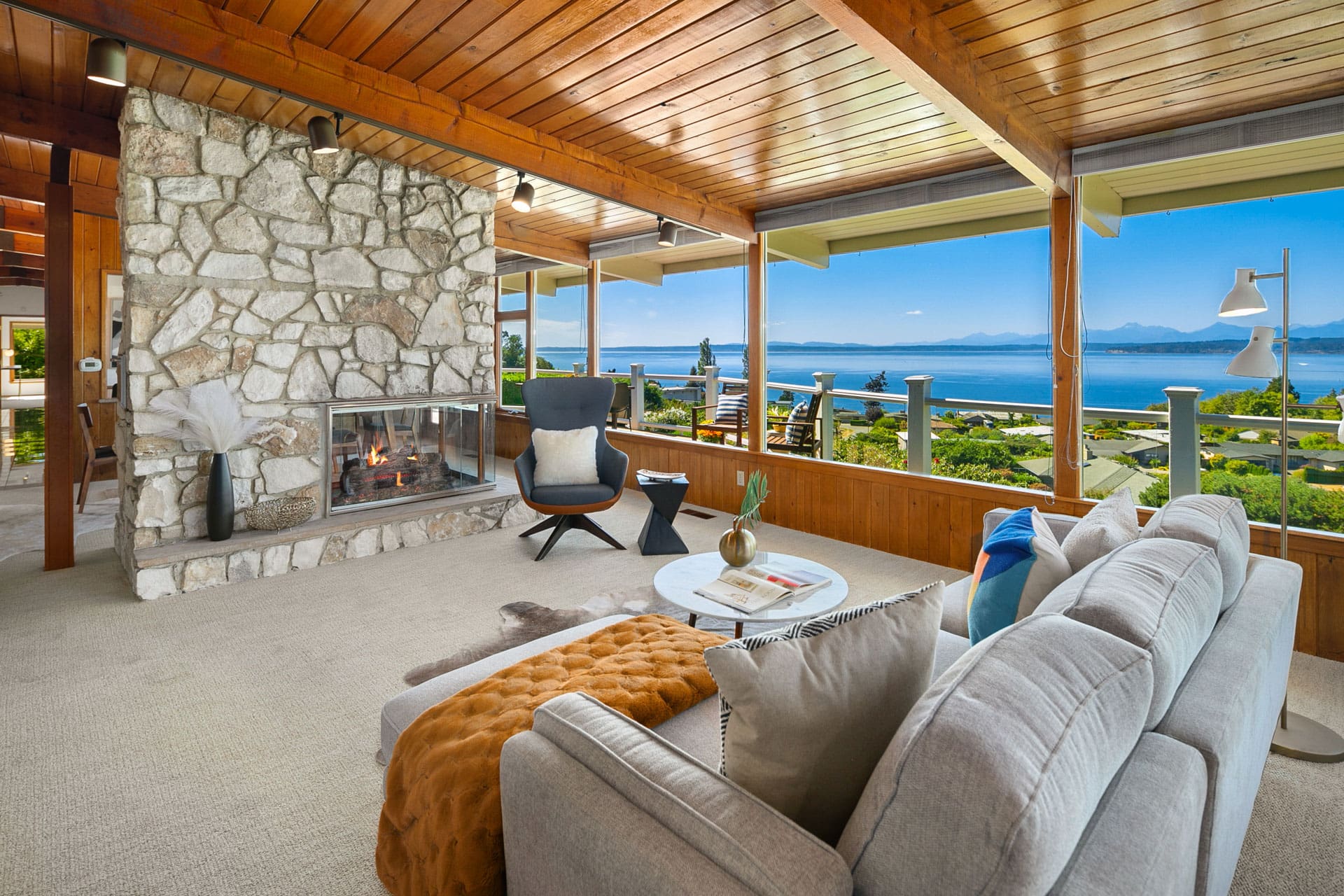 A cozy living room with a large stone fireplace and a modern gray couch. The room features wooden ceilings and large windows offering a panoramic view of the ocean and mountains. There's a black chair, side table, and a shaggy brown throw blanket on the couch.