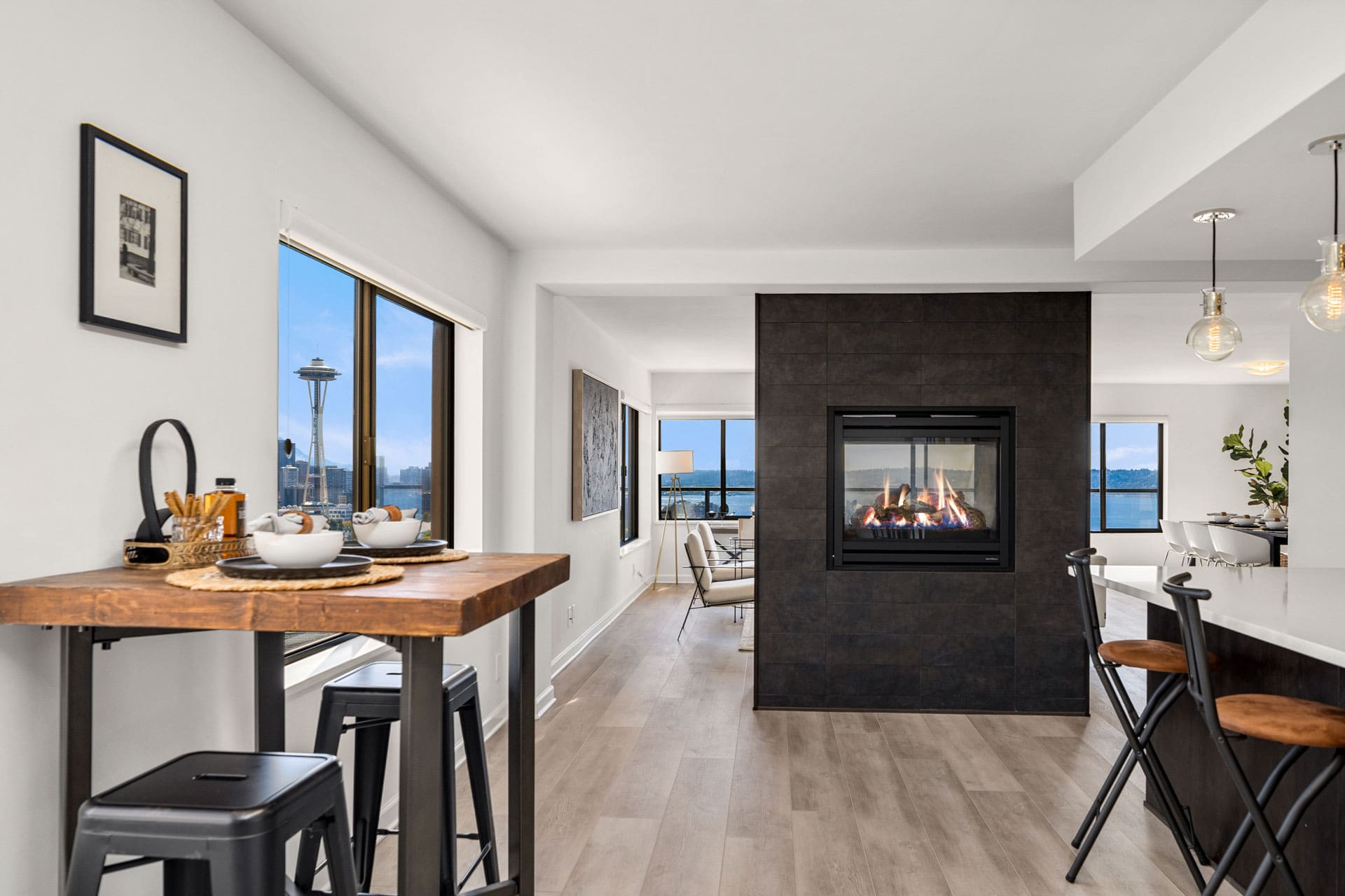 Modern apartment interior with an open floor plan, featuring a wooden dining table with barstool seating, a central dual-sided fireplace, and large windows showcasing a view of the city, including the Space Needle. The space is bright and stylishly furnished.