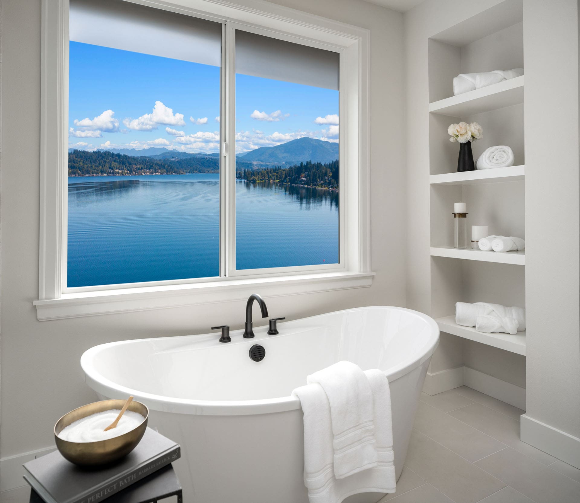 Modern bathroom with a white freestanding bathtub in front of a large window offering a view of a serene lake and mountains. Shelves on the right hold towels and decorative items. Neutral tones and clean design create a calming atmosphere.