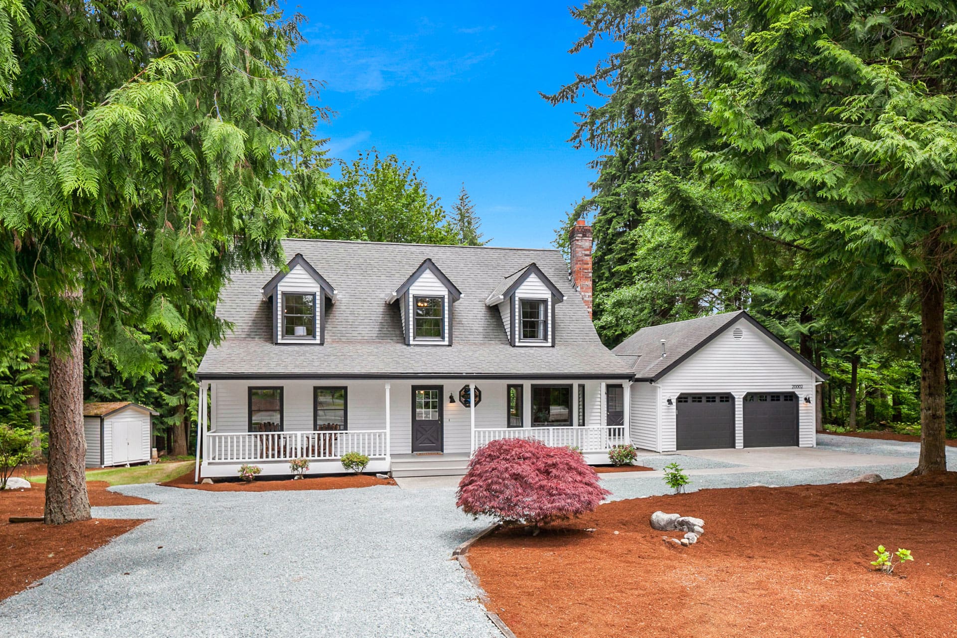 A charming white two-story house with a gray roof sits amidst lush greenery. It features a covered porch, dormer windows, a gravel driveway leading to a two-car garage, and a well-maintained yard with a small tree in front. Tall trees surround the property.