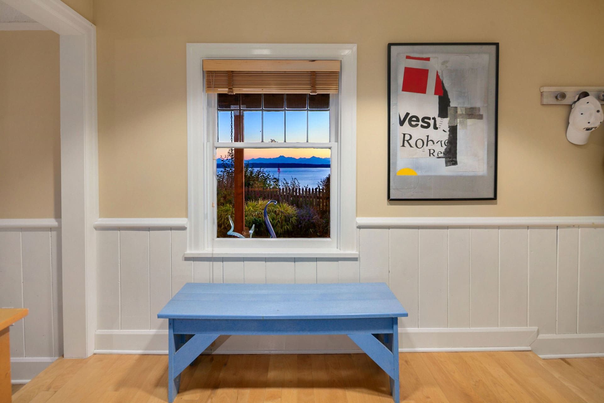 A room with light-colored walls and wainscoting features a blue table in front of a window. Through the window, a scenic view of a body of water and mountains can be seen. On the wall to the right, there's a framed abstract artwork and a coat hanger with hats.