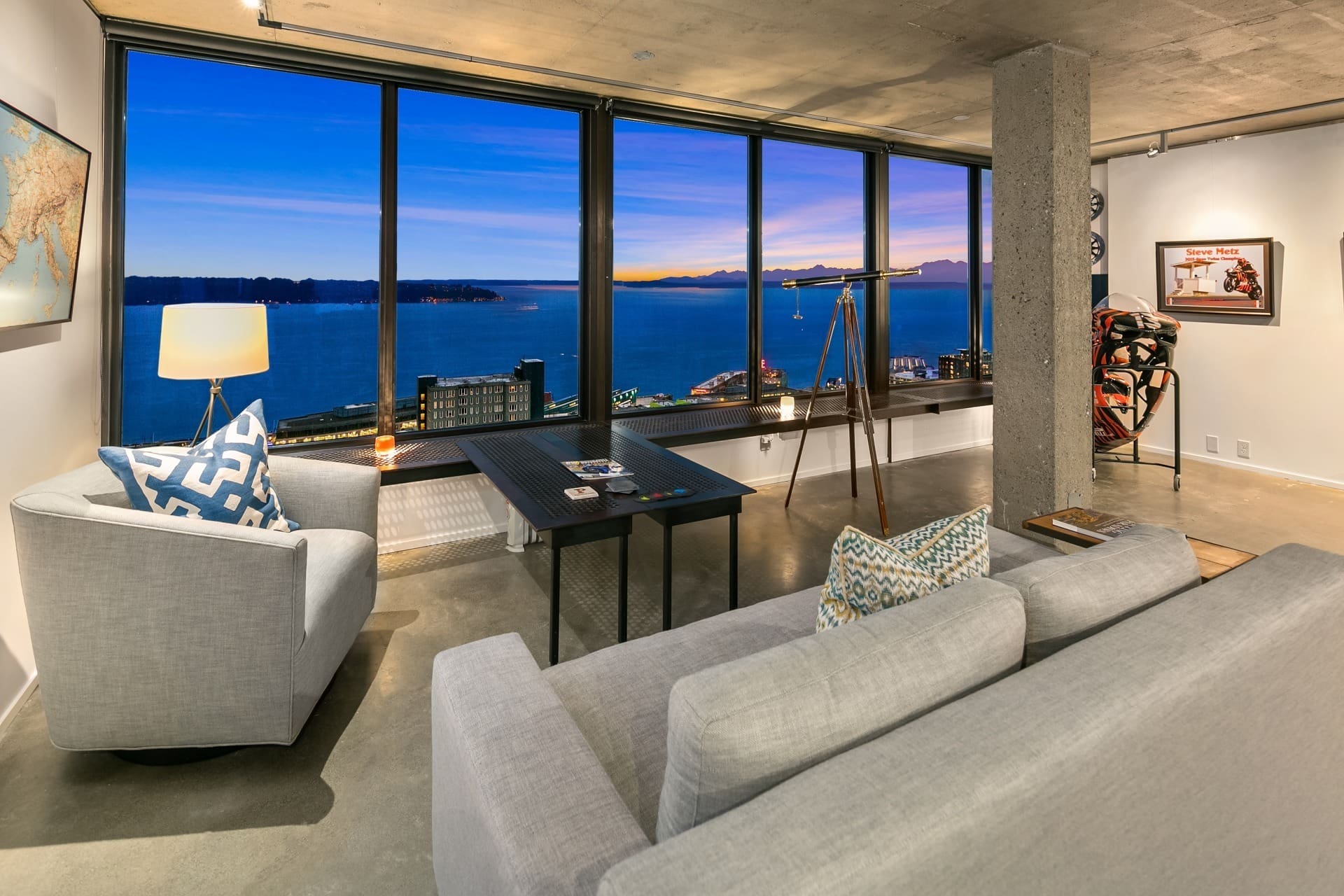 A modern living room with large windows offering a panoramic view of a cityscape and ocean at sunset. The room features a gray sectional sofa, a dark wooden table, a telescope by the windows, and wall art, including a framed sports jersey and vintage poster.