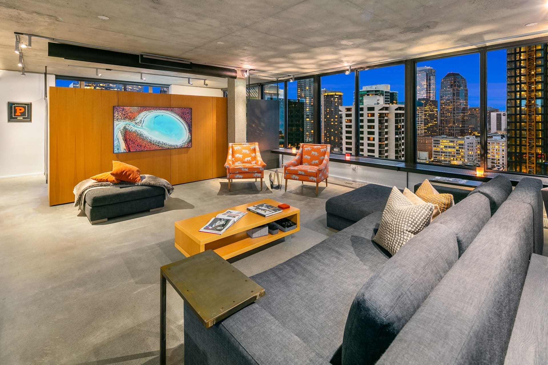 Modern living room with floor-to-ceiling windows showcasing a city skyline view. The room features a large gray sectional sofa, two orange patterned armchairs, a wooden coffee table, and a contemporary painting mounted on a partition wall.