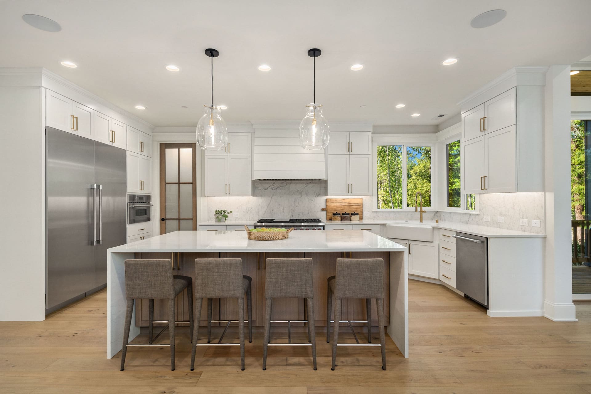 Modern kitchen with white cabinetry and a large central island with a white countertop. The island has four gray upholstered barstools. Stainless steel appliances, two pendant lights, and a window with a view of greenery complete the space.