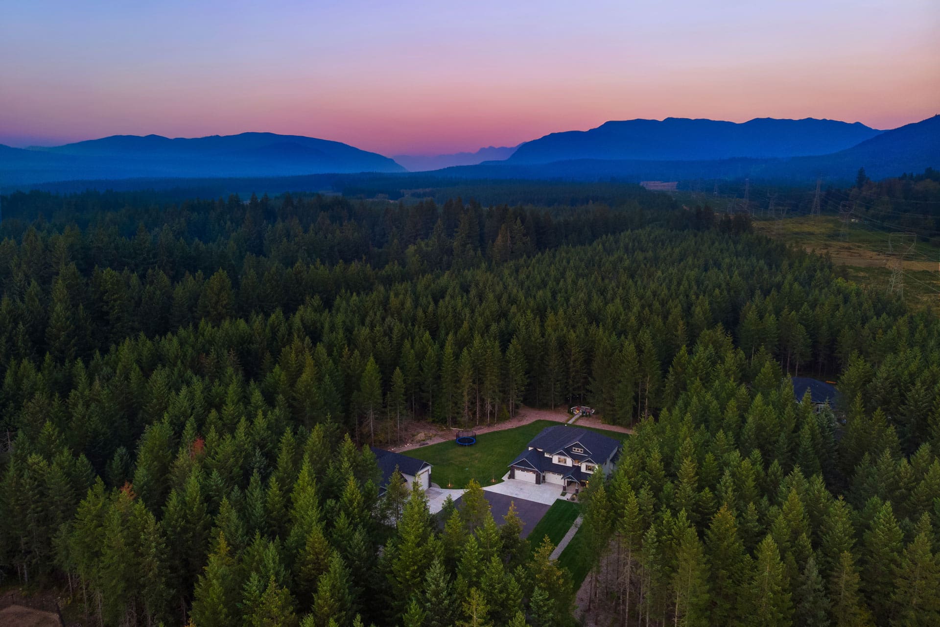 Aerial view of a secluded house surrounded by dense forest at dusk. The sky displays a gradient of colors from pink to deep blue as the sun sets. Mountains are visible in the background, fading into the horizon.