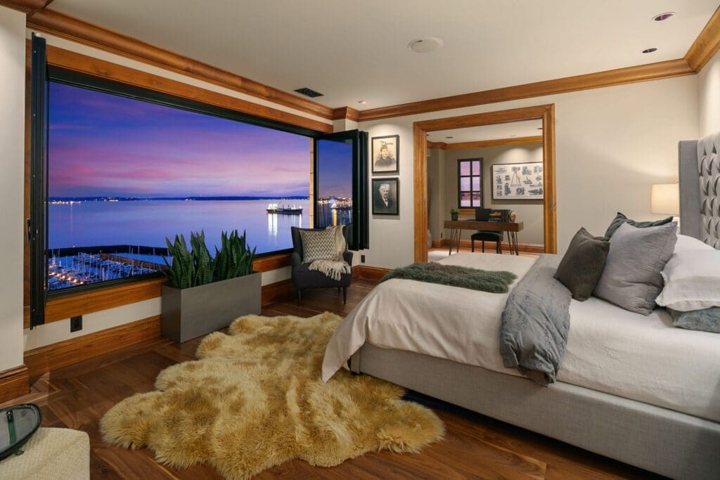 A cozy bedroom with a large open window facing a peaceful harbor at sunset. The room features a plush rug, modern furniture with grey and white bedding, potted plants, and wooden accents, creating a warm and inviting atmosphere. Photos and pictures adorn the walls.