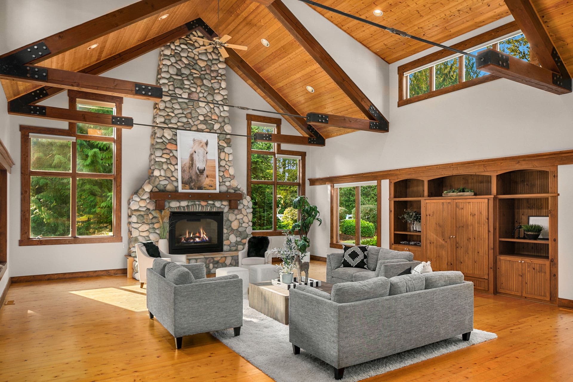 A spacious living room with vaulted wooden ceilings and exposed beams. A large stone fireplace with a horse painting above it serves as the focal point. Grey sofas and a wooden coffee table sit on a light rug. Tall windows bring in natural light and views of greenery.