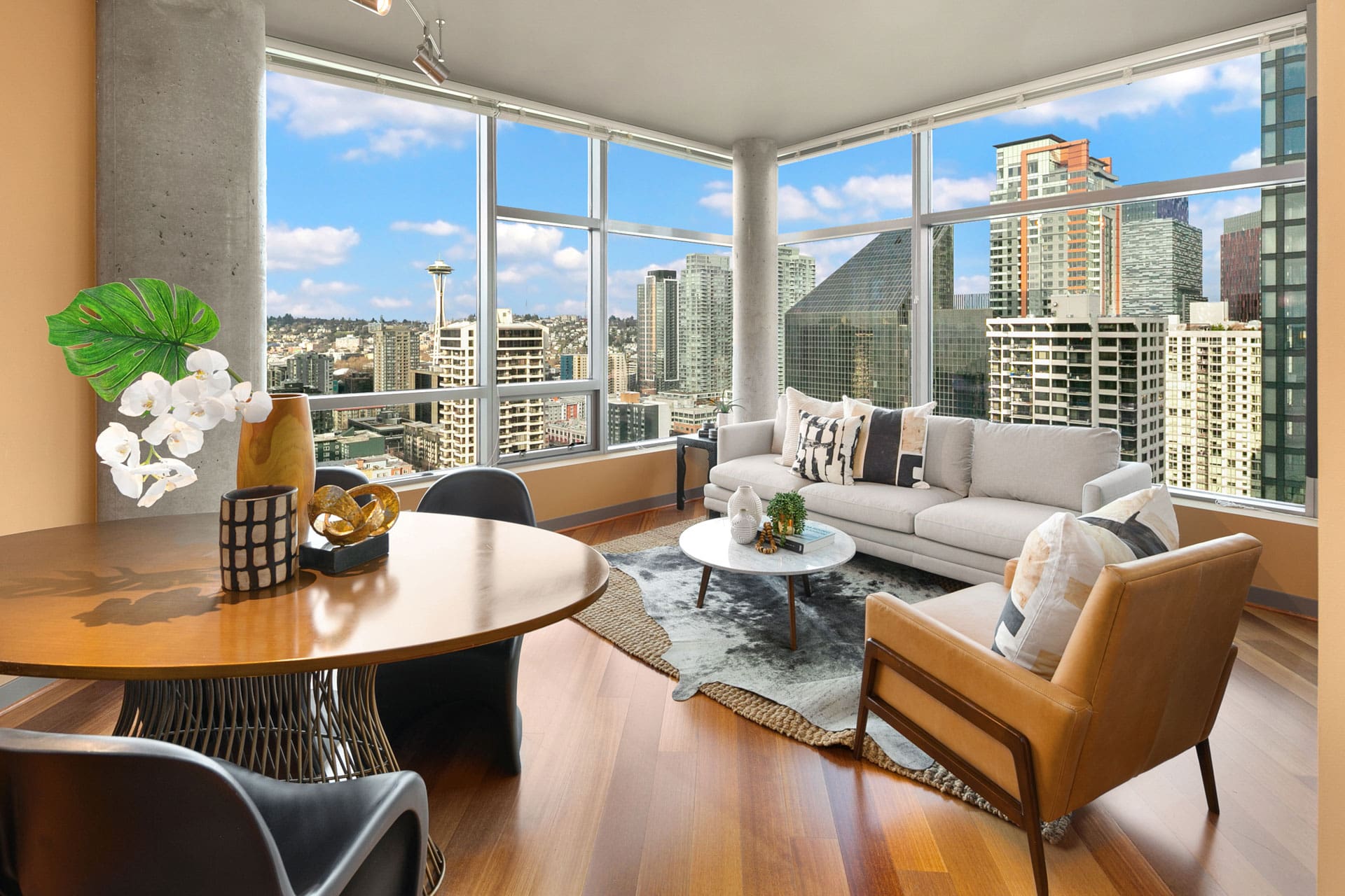 A modern living room with floor-to-ceiling windows, offering a panoramic city view. The space features a beige sectional sofa, a coffee table, an armchair, a round dining table with chairs, and elegant decor including a vase with white flowers.