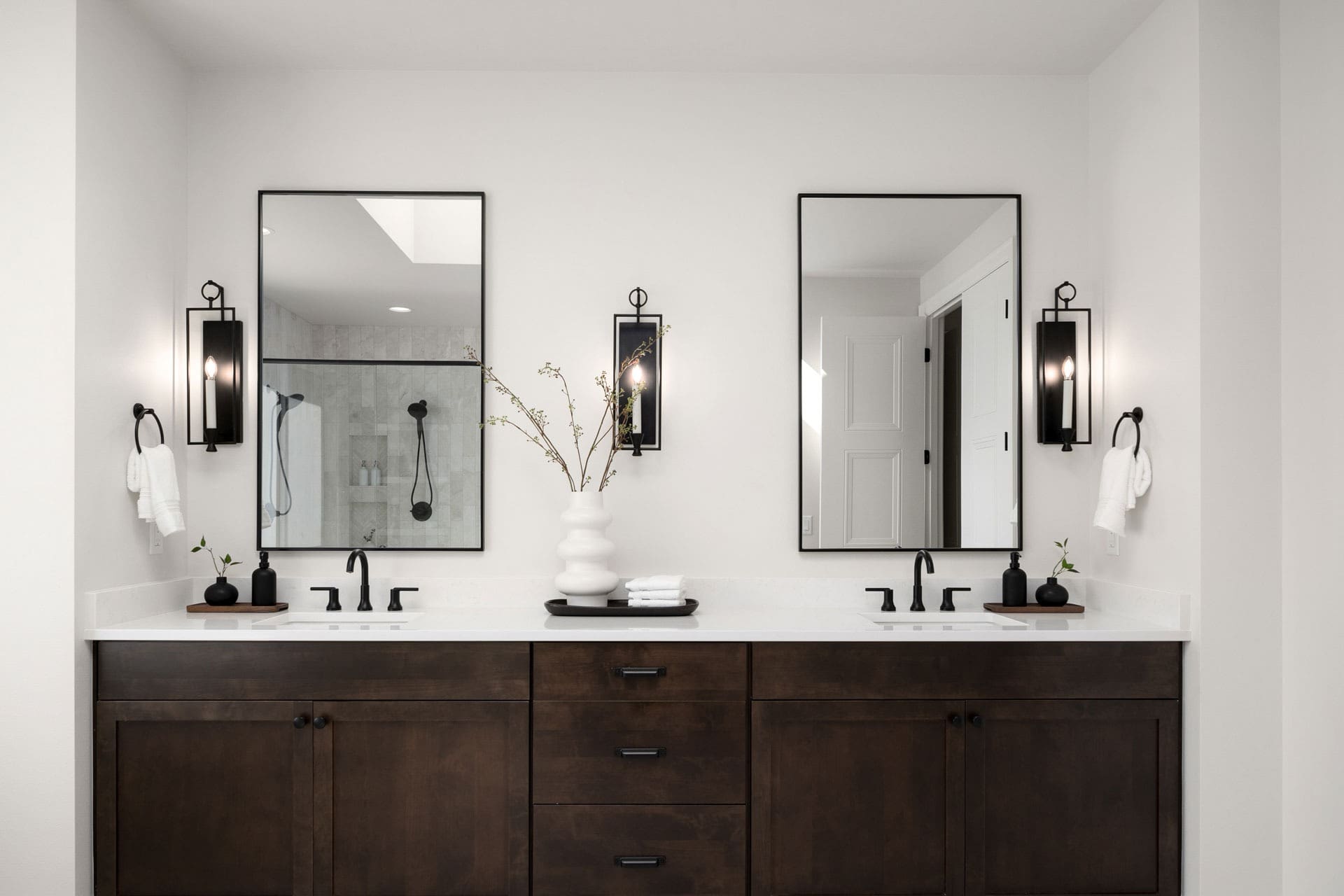 A modern bathroom features a double vanity with dark wooden cabinets, white countertops, and black faucets. Above the sinks are two rectangular mirrors with black frames, each flanked by black wall sconces. The background shows a shower with a glass door.