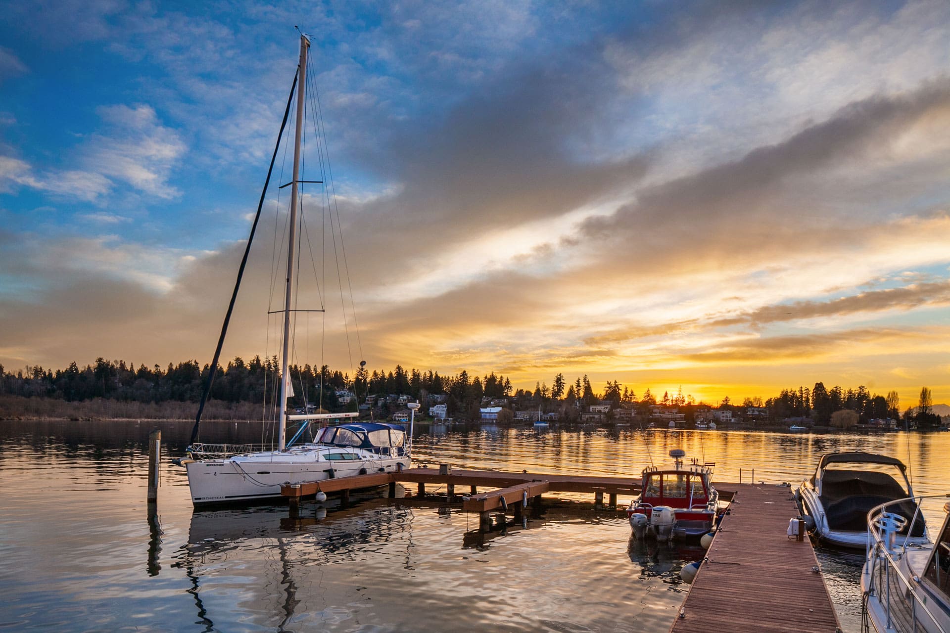 A serene waterfront scene at sunset with boats docked at a wooden pier. The sky is partly cloudy with hues of orange and yellow, reflecting on the calm water. Trees and houses line the shore in the background, completing the tranquil evening setting.