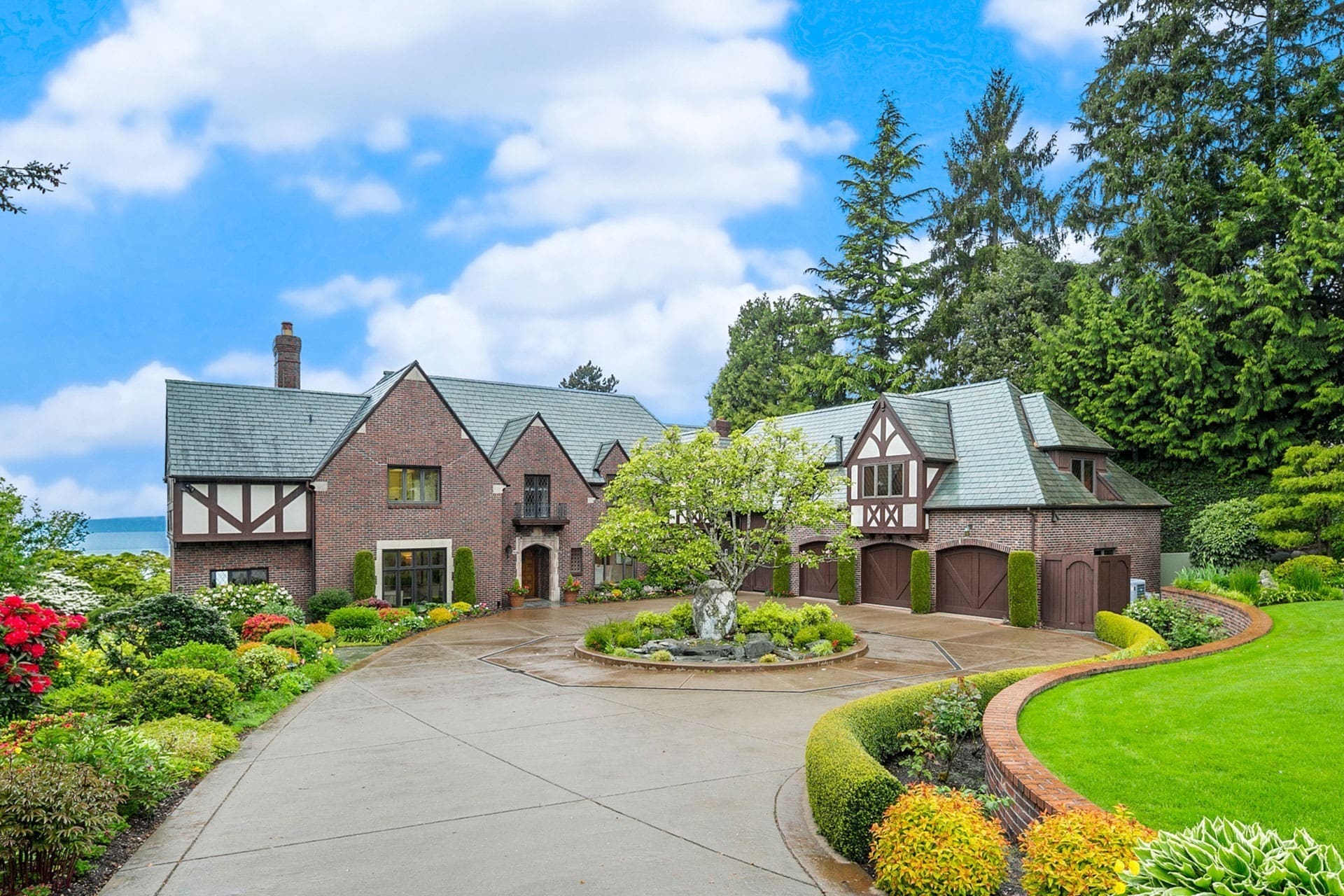 A large, picturesque Tudor-style house with brick exterior, multiple gables, and wooden accents. It is surrounded by lush greenery, colorful flowers, and manicured lawns. A circular driveway leads to the entrance with garages visible to the right. The sky is partly cloudy.