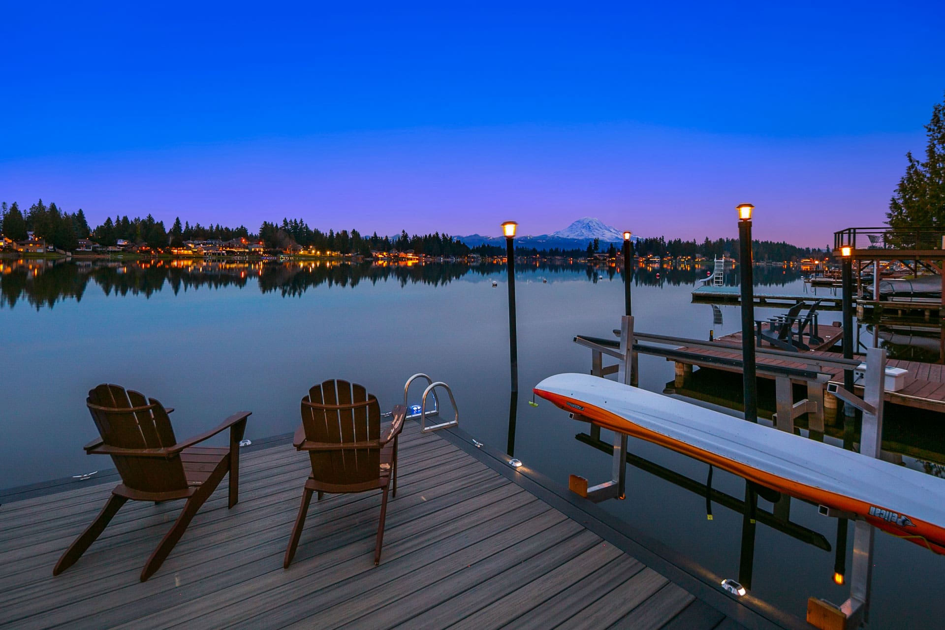 A serene lakeside scene at dusk, featuring two wooden chairs on a dock facing calm water. A canoe is stored on the right side of the dock, with softly glowing lights illuminating the scene. Trees and houses line the shore, with a snow-capped mountain visible in the distance.