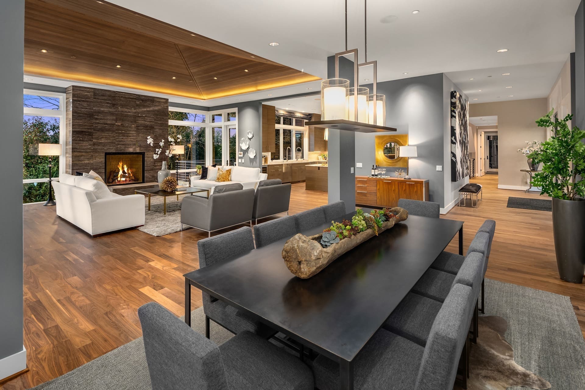 A contemporary open-plan living and dining area features hardwood flooring, a stylish wood-stone fireplace, grey and white sofas, and a sleek black dining table with grey chairs. Modern pendant lights and a large wooden centerpiece highlight the table.