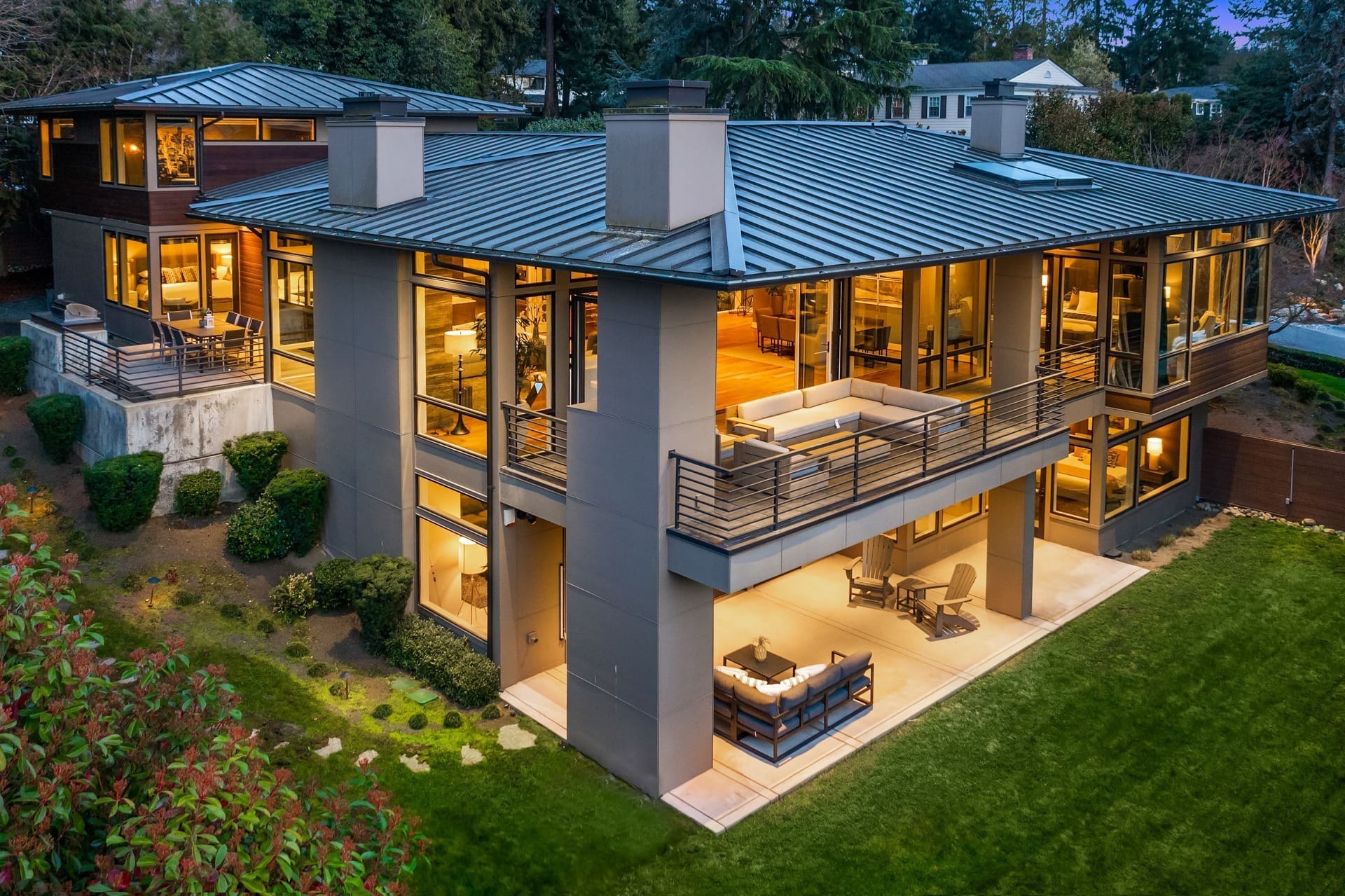 A modern, two-story house with large windows and a flat roof, illuminated warmly from the inside. The exterior features a spacious outdoor patio with seating areas on the lower and upper levels. The surrounding landscape includes well-maintained lawns and shrubs.