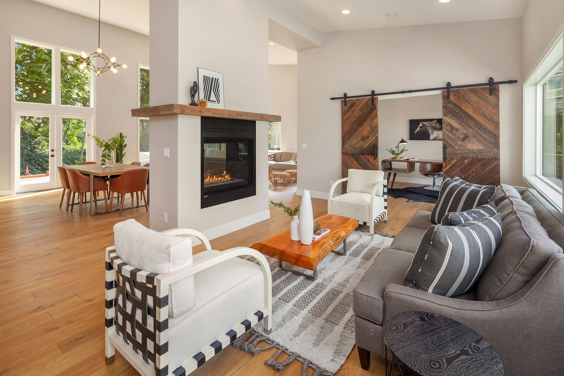 Modern living room with a cozy seating area featuring a gray sofa and two patterned armchairs around a wooden coffee table. A fireplace separates the living room from a dining area with large windows. Sliding barn doors lead to a study area with a desk and chair.