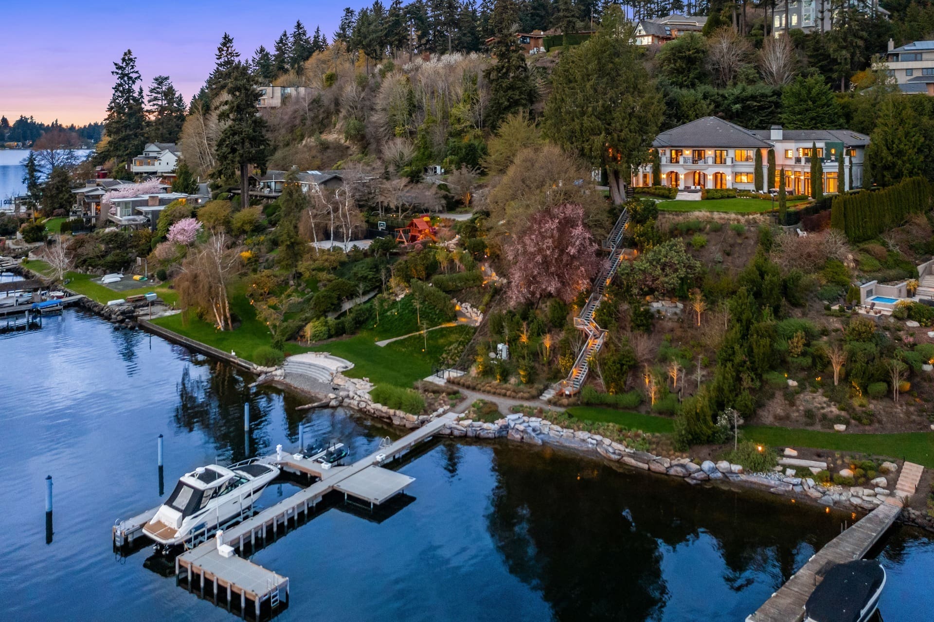 Aerial view of a picturesque lakeside estate at dusk. The scene features luxurious houses nestled among trees on a hillside, a well-lit mansion at the top, and private docks with boats along the tranquil water's edge. The sky transitions to a soft purple hue.