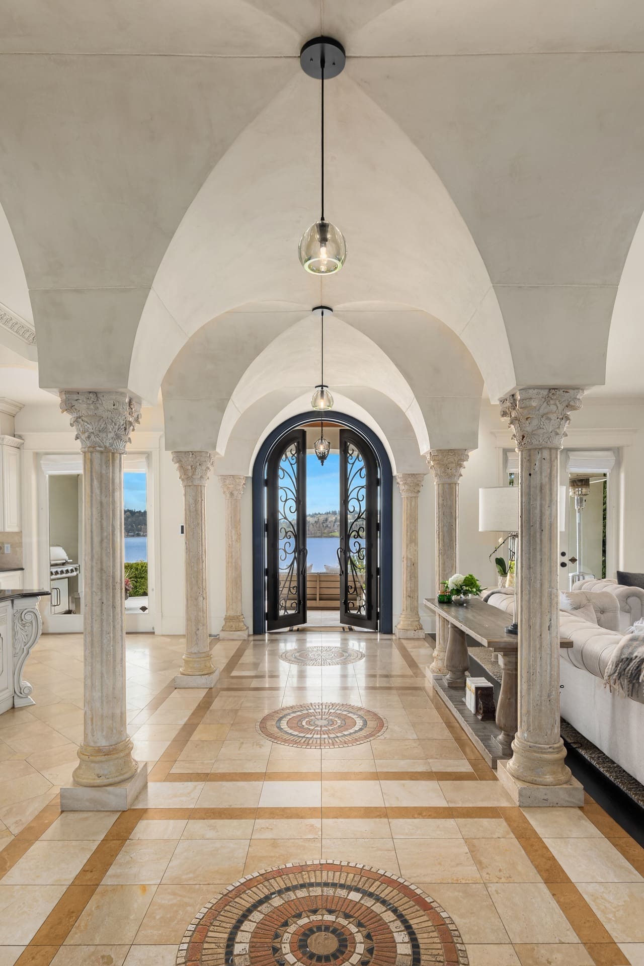 A luxurious hallway with arched ceilings and ornate columns leads to a double door with decorative ironwork. The tile floor features intricate circular patterns. To the right, a living area is partially visible with a sofa, lamp, and an arrangement of plants.