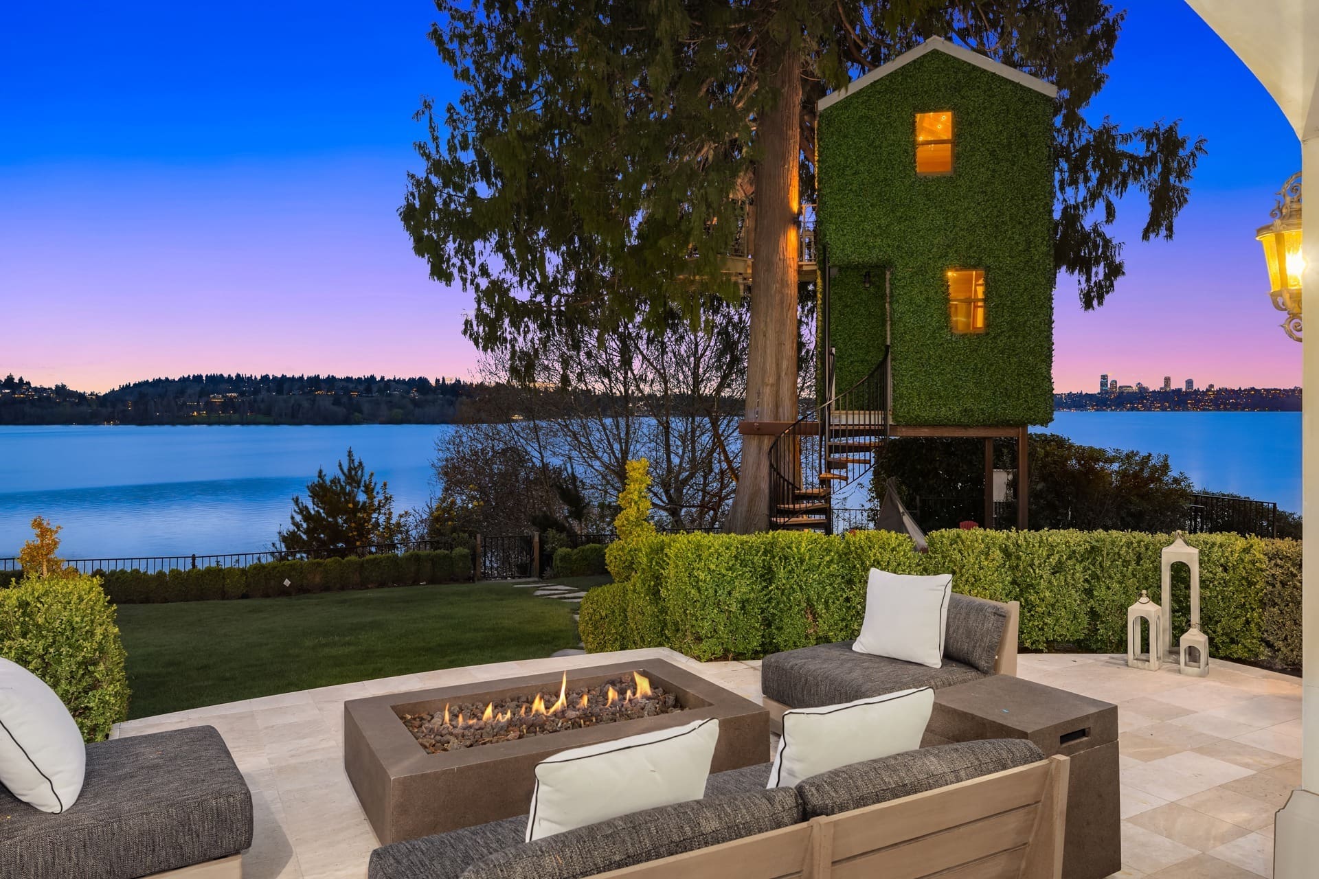 A serene lakeside patio with a fire pit and cushioned seating in the foreground. In the background, a lush green treehouse is nestled in a large tree. The scene overlooks a calm lake with rolling hills and a city skyline in the distance at dusk.