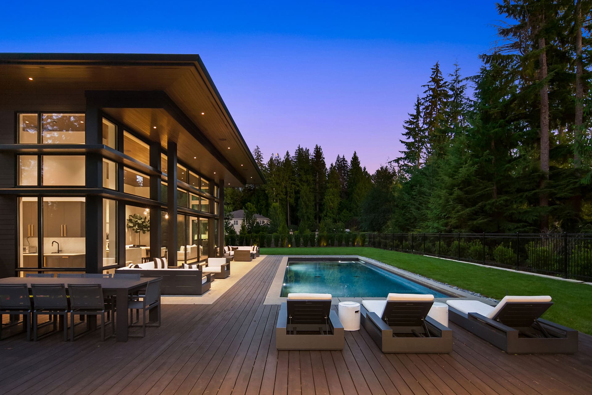 A modern home with large glass windows and a sloped roof at twilight. The backyard features a spacious wooden deck with outdoor seating and a dining area, adjacent to a rectangular swimming pool surrounded by lounge chairs. The background shows a forested area.