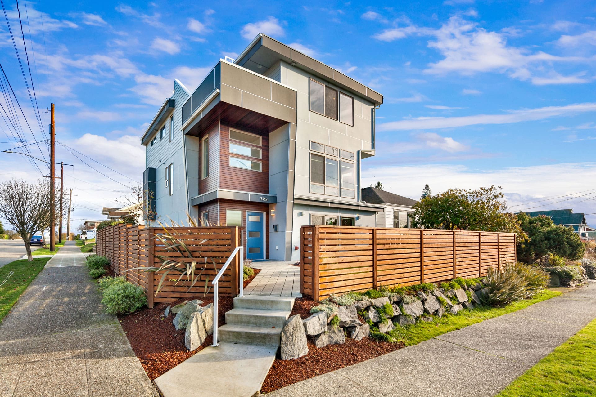 A modern, multi-story house with large windows and a mix of grey and wooden exterior panels. It features a pathway leading to a blue front door, a landscaped front yard with rocks and greenery, and a wooden fence enclosing the property on a corner lot under a bright blue sky.