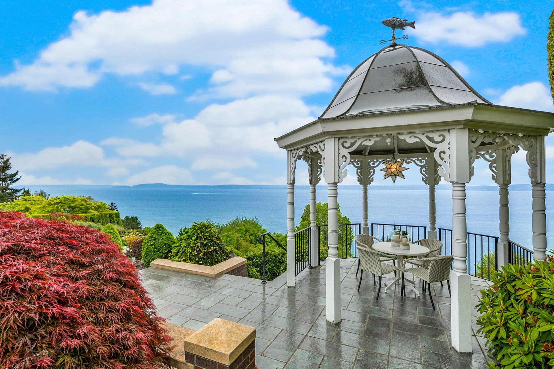 A white gazebo with ornate trim stands on a tiled patio overlooking the ocean. Inside the gazebo is a round table with four chairs. Surrounding the area are lush green bushes, a red-leafed plant, and trees. A partly cloudy sky is above the tranquil seascape.