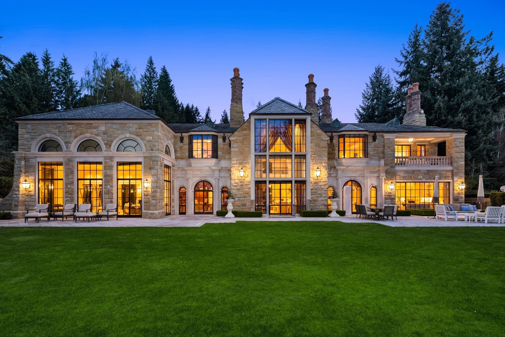 A large, luxurious two-story mansion with numerous tall, arched windows illuminated warmly from within. The exterior features stone facade and towering chimneys. In the foreground is a well-manicured green lawn, surrounded by trees at dusk.