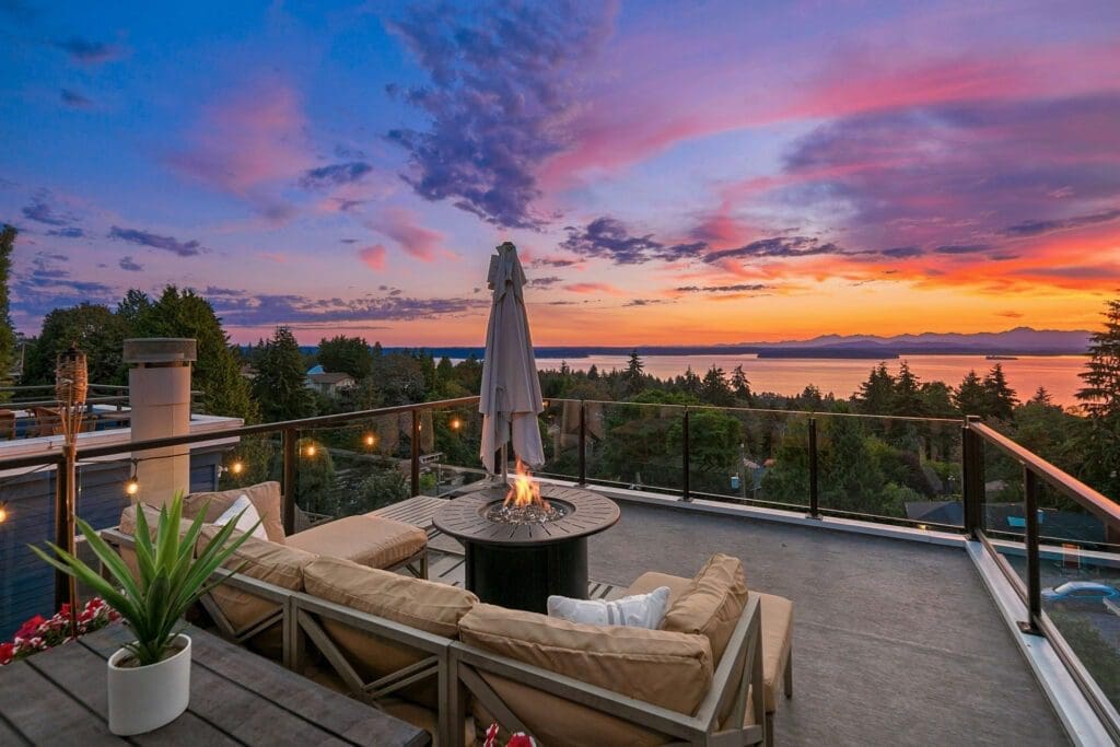 A cozy outdoor patio overlooking a serene landscape with trees and a body of water at sunset. The scene features cushioned seating around a fire pit table, a closed umbrella, and string lights, creating a warm and inviting atmosphere. The sky is vibrant with pink, purple, and orange hues.