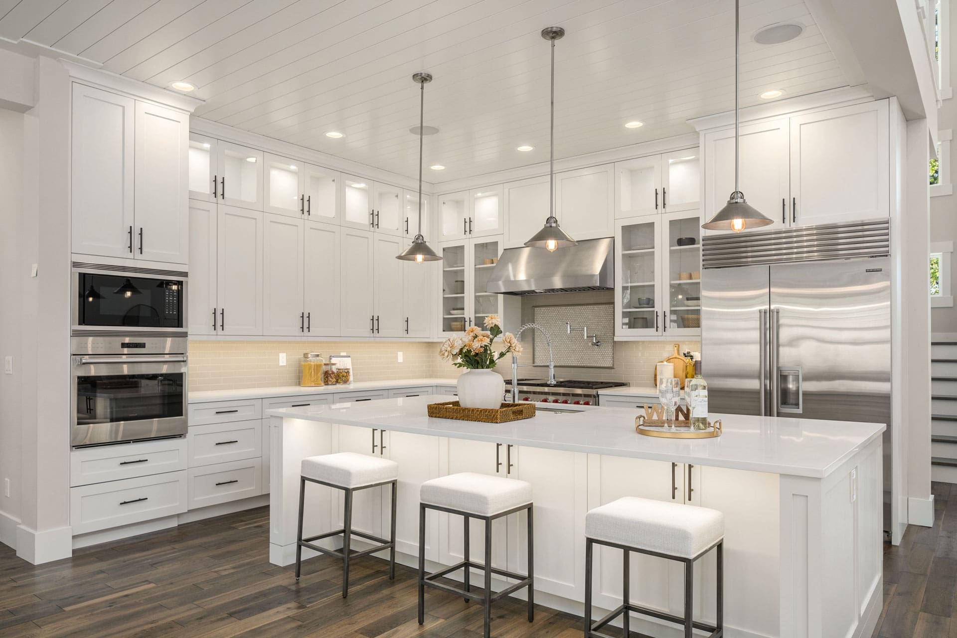 A modern kitchen features white cabinetry, stainless steel appliances, and a large island with white countertops. Pendant lights hang above the island, which is adorned with a vase of flowers and a tray. Three cushioned stools line the island, and the floor is dark wood.