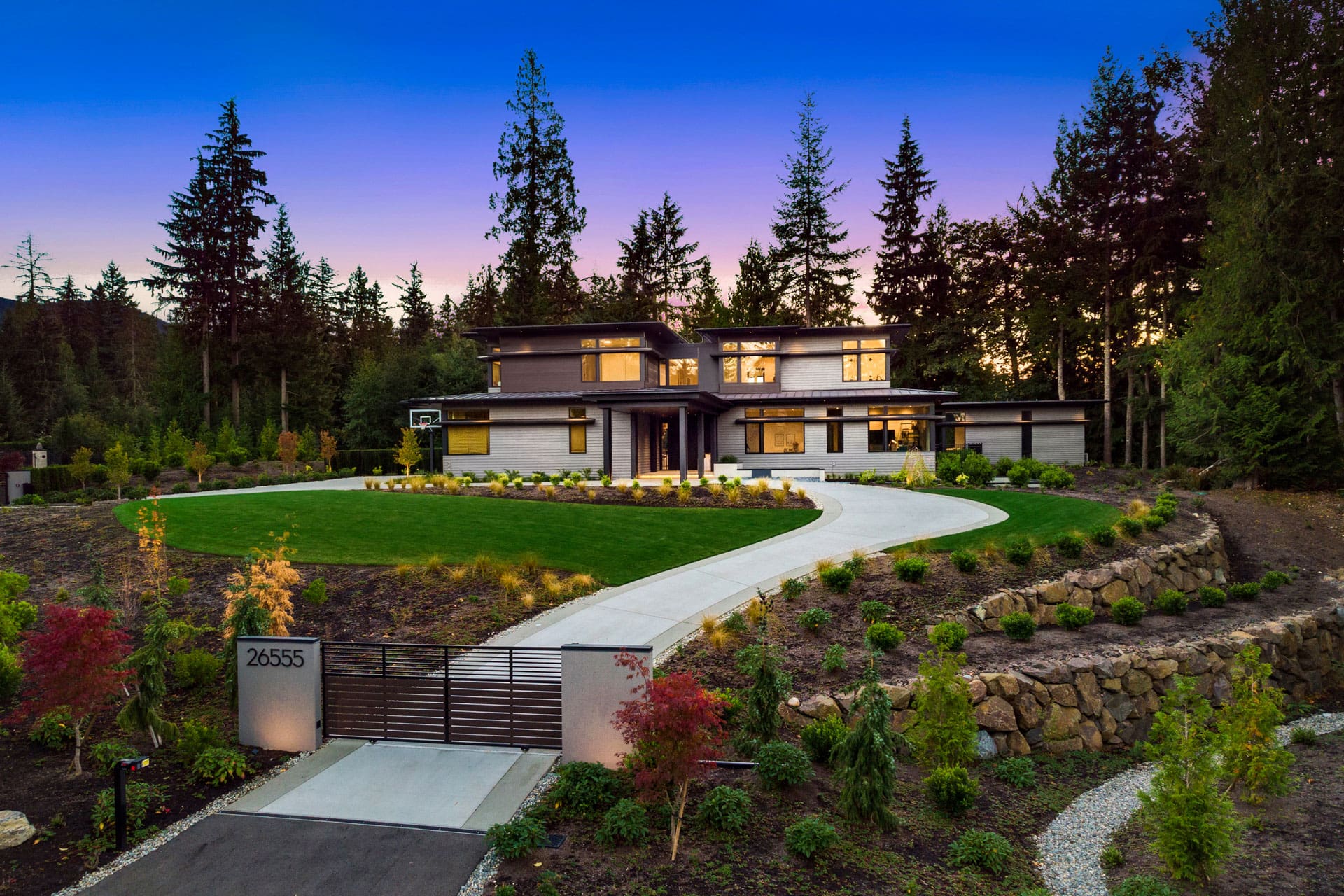 A modern two-story house with large windows is surrounded by trees and landscaping. A winding driveway leads to the front entrance, and a manicured lawn is situated in front. The address "26555" is displayed on a gate at the driveway's entrance, with a sunset sky behind.