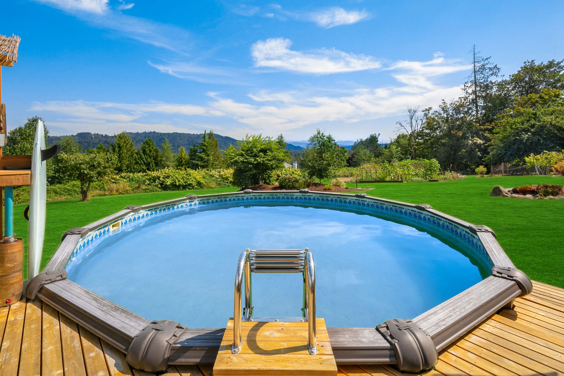 A round, above-ground swimming pool with metal ladder on a wooden deck is surrounded by lush green grass. In the background, there are trees and rolling hills under a blue sky with scattered clouds.