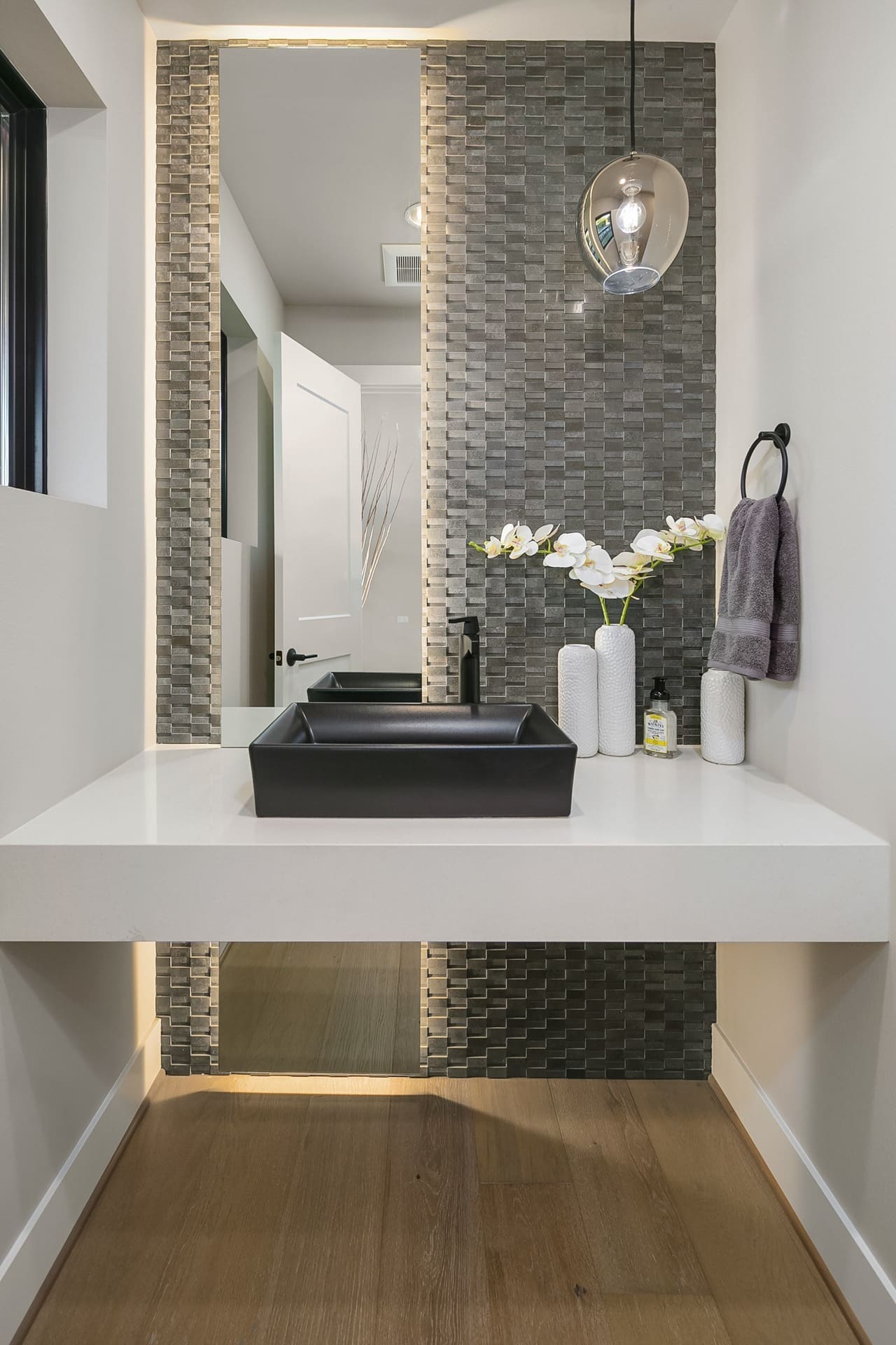 Modern bathroom with a floating white vanity featuring a black rectangular sink. A textured gray tiled backsplash extends from the counter to the ceiling, accented by a round glass pendant light. The space is accented with white vases and towels on a black ring.