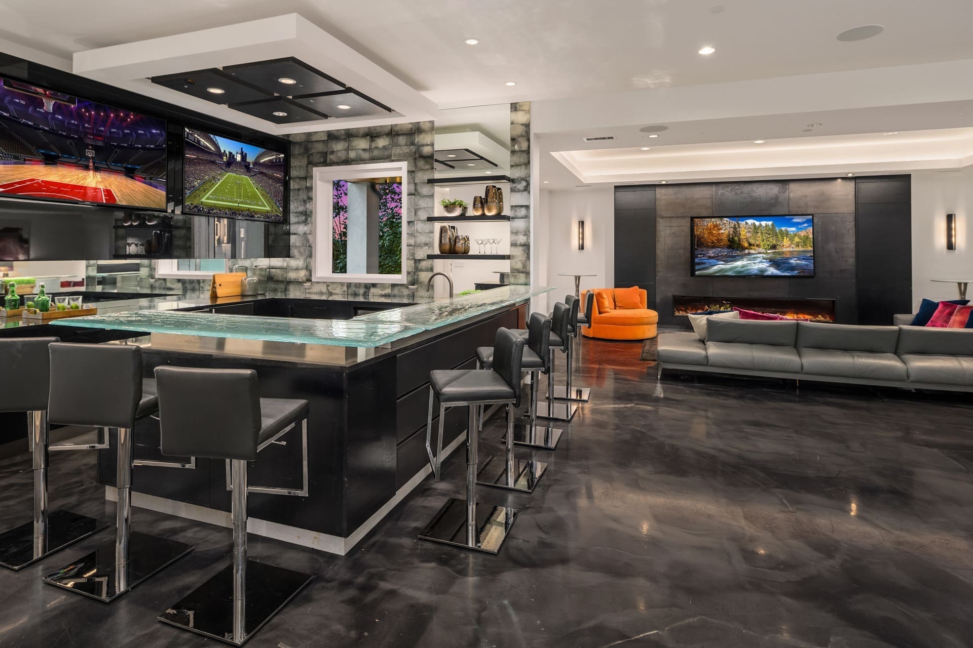 A modern entertainment room with a sleek bar area, featuring high-top chairs and three mounted flat-screen TVs. An adjacent lounging area has a gray couch, an orange chair, and a large TV above a fireplace. The room has a polished dark floor and elegant decor.