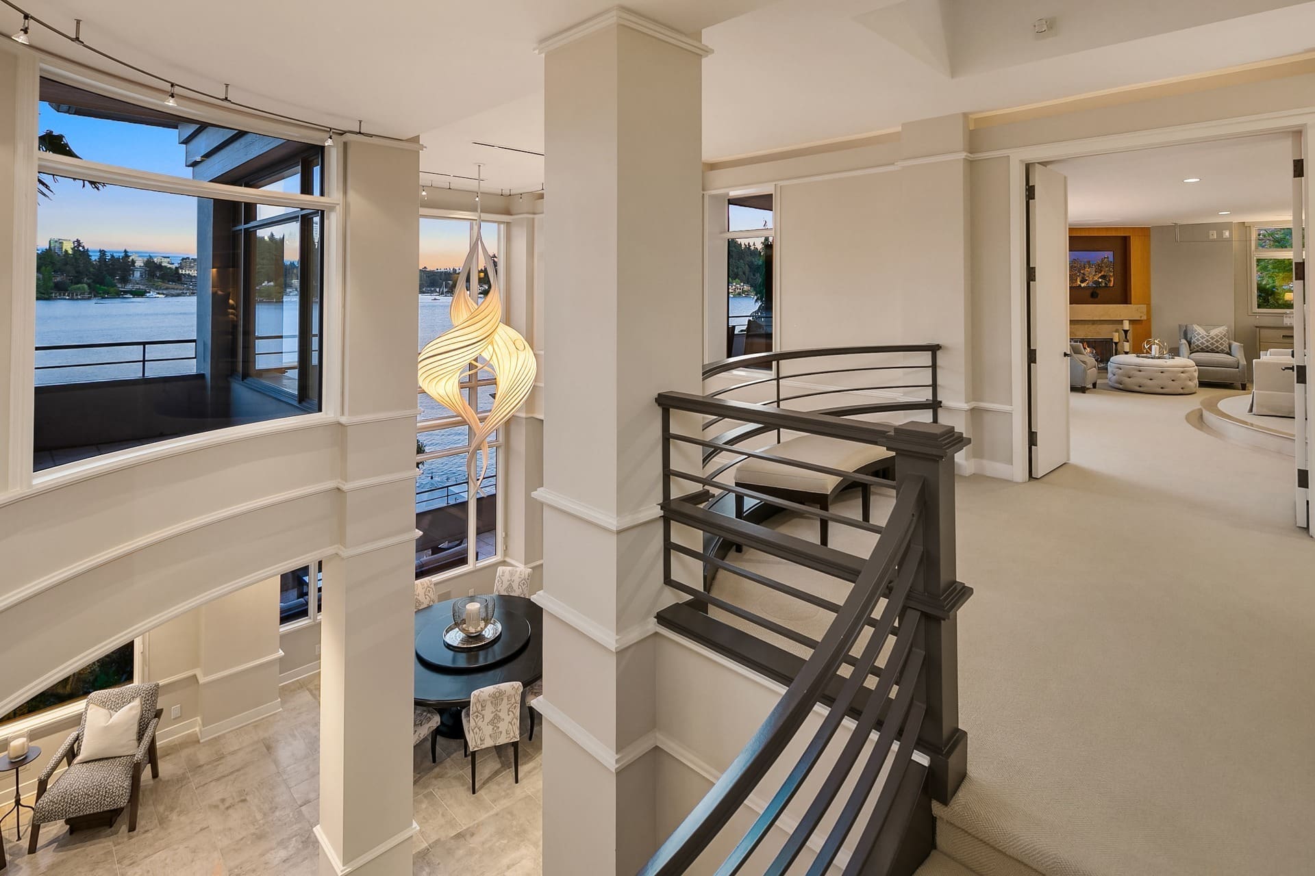 A modern, spacious home interior with a view of a lake through large windows. A staircase with dark railings leads from the lower dining area to an upper level. A unique pendant light hangs above the dining table set with chairs. The upper level features a cozy living area.