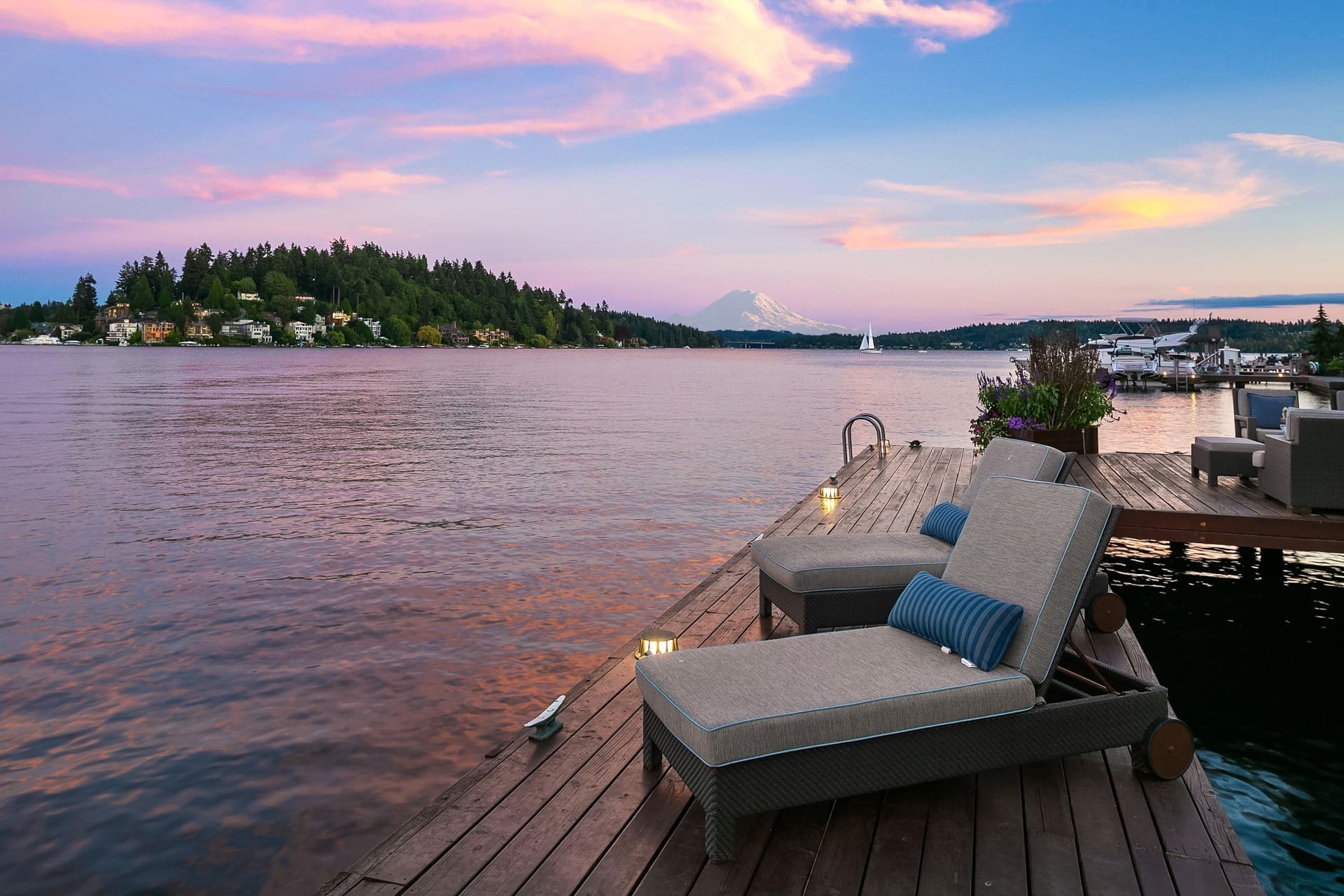 A serene lakeside scene at sunset, featuring two lounge chairs on a wooden dock. The water reflects the pink and blue hues of the sky, with an island and distant mountain visible. Boats are docked nearby, complementing the tranquil atmosphere.