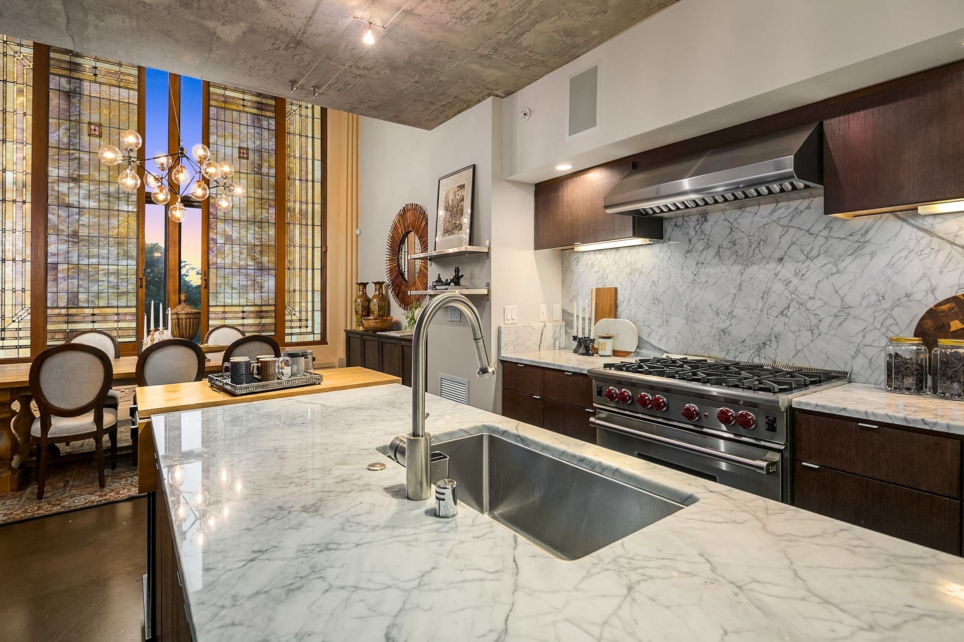 Modern kitchen with marble countertops, stainless steel appliances, gas range, and sleek dark wood cabinetry. Dining area with a wooden table and upholstered chairs is visible in the background, featuring tall windows with decorative, geometric patterned glass.