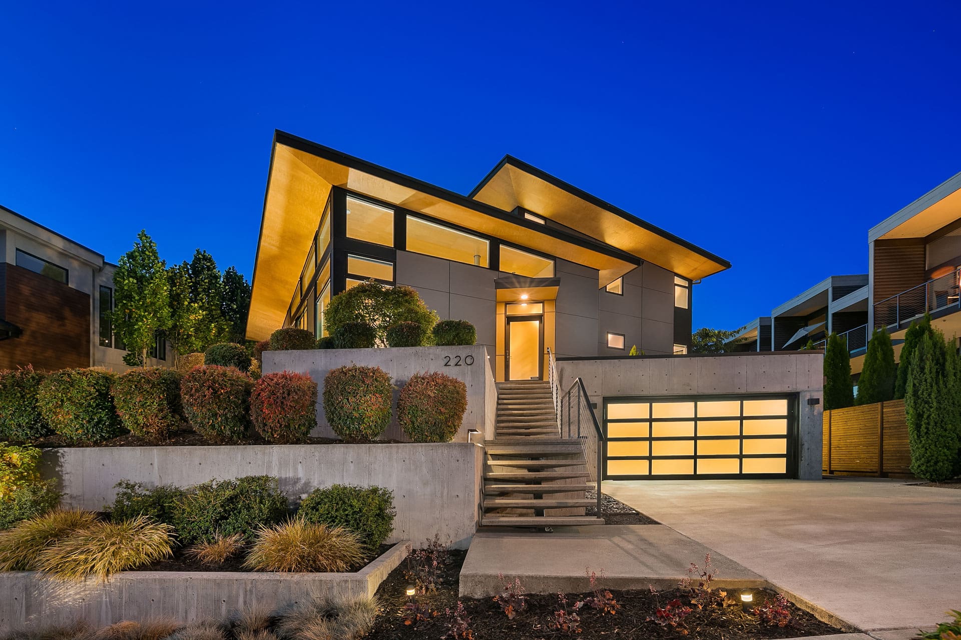 A modern house with angular rooflines illuminated at dusk, featuring large windows and a mix of wood and concrete exterior. The property has a landscaped front yard with steps leading to the entrance, a double garage, and well-maintained shrubs and bushes.