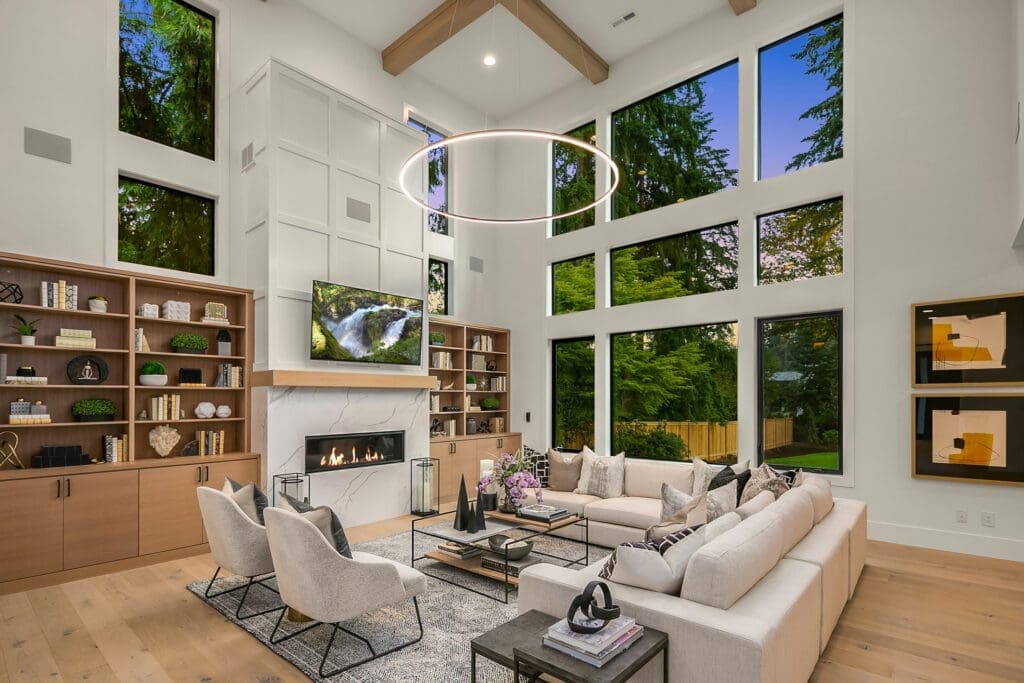 A spacious, modern living room with large windows offering a forest view. The room features a high ceiling with wooden beams, a chandelier, a fireplace, and a TV above it. There's a U-shaped sectional sofa, two armchairs, and two bookshelves flanking the fireplace.