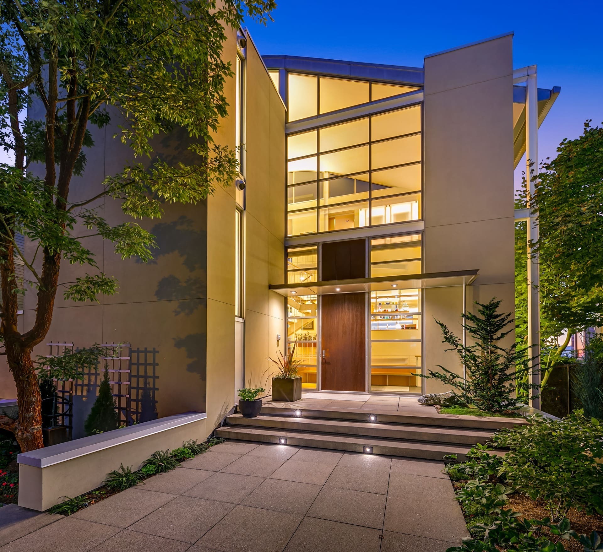 A modern, two-story house entrance is shown at dusk. The house features large, multi-pane windows and a minimalist design with clean lines. Steps lead up to a wooden front door. Trees and plants surround the walkway, enhancing the contemporary aesthetic.