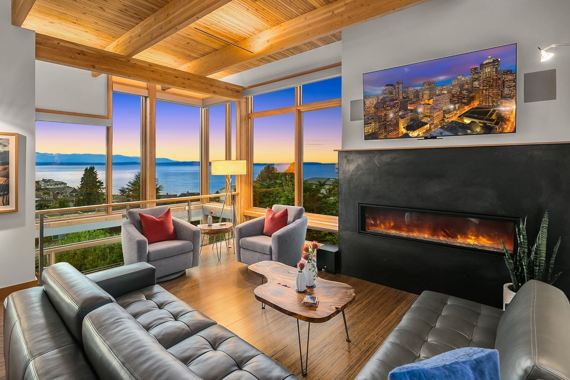 A modern living room with a wall-mounted fireplace under a large TV. There's a cityscape and ocean view visible through large, expansive windows. The room features wooden beams, two grey armchairs, a wooden coffee table, and a grey sectional sofa.