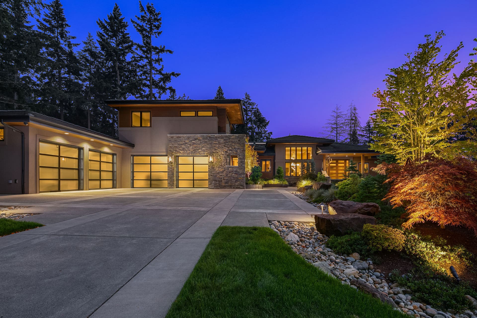 Modern two-story house with large windows and garage doors, illuminated at dusk. The exterior features a mix of stone and plaster finishes, surrounded by a well-manicured yard with lush greenery and colorful trees.