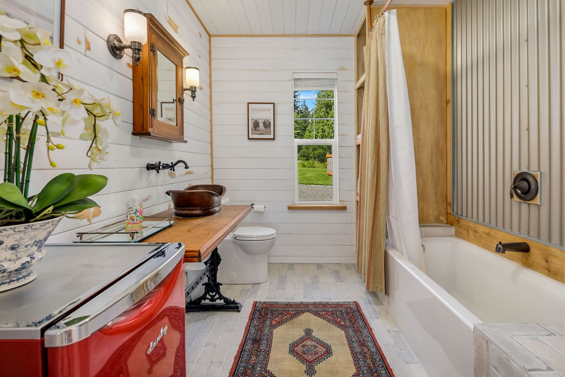 A bright bathroom with white shiplap walls, a red mini-fridge, a rustic wooden vanity with a vessel sink, a toilet, and a bathtub with a curtain. There's a vintage rug on the floor and a window with a view of a green garden. A potted orchid sits on the countertop.