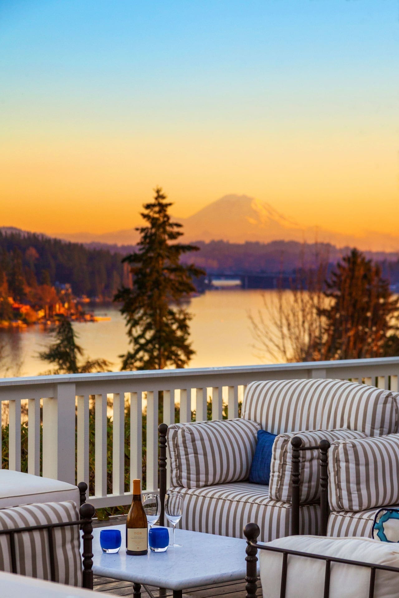 A cozy outdoor seating area with striped cushions overlooks a tranquil lake at sunset. A mountain is visible in the distance. On a table, there is a wine bottle, wine glasses, and two blue cups. The sky is painted in gradient hues of orange and yellow.