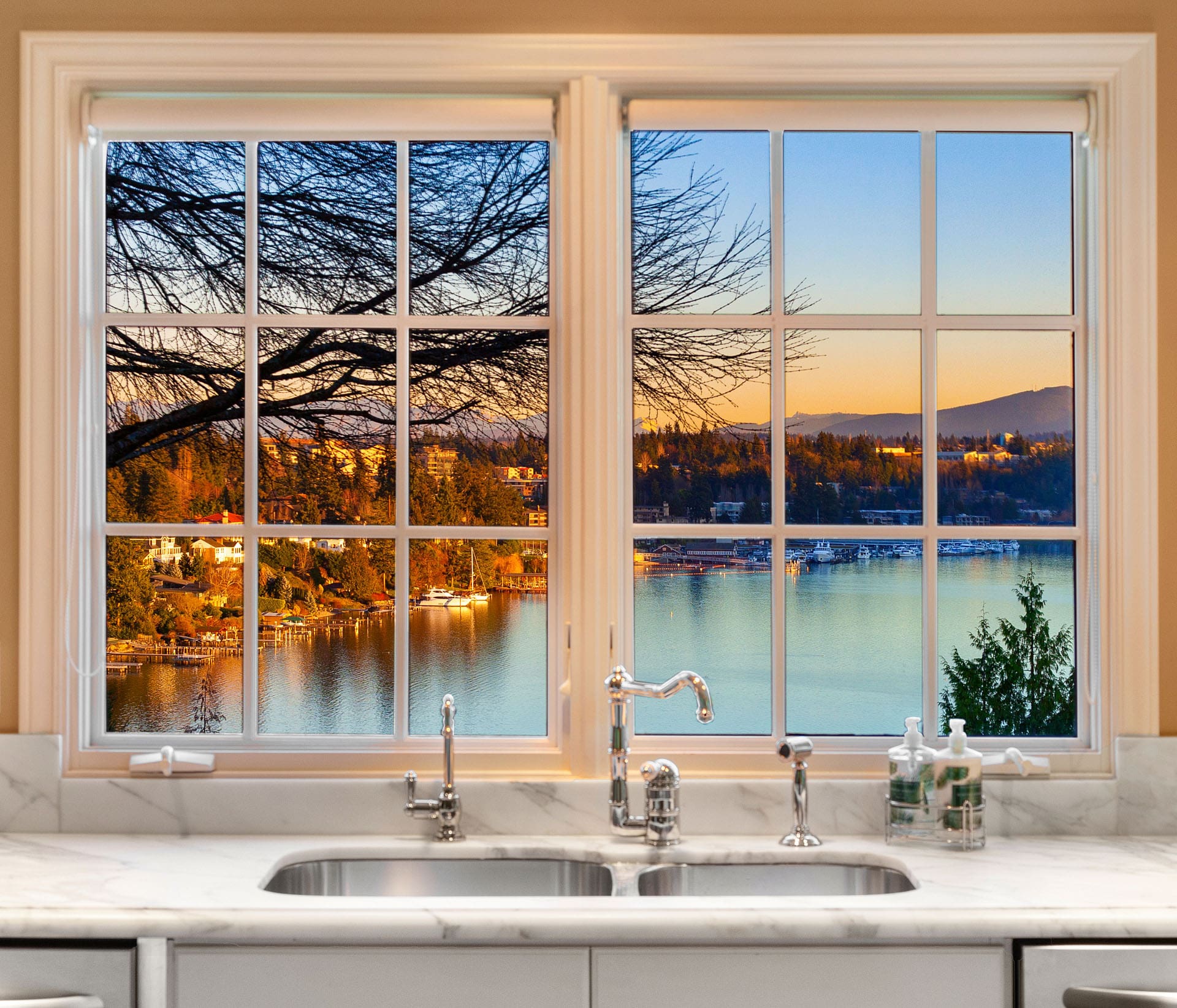 A kitchen window with a white frame overlooks a stunning lake surrounded by autumn foliage. The view includes a bright horizon with mountains in the distance and houses lining the shore. The kitchen sink and faucets are visible below the window.