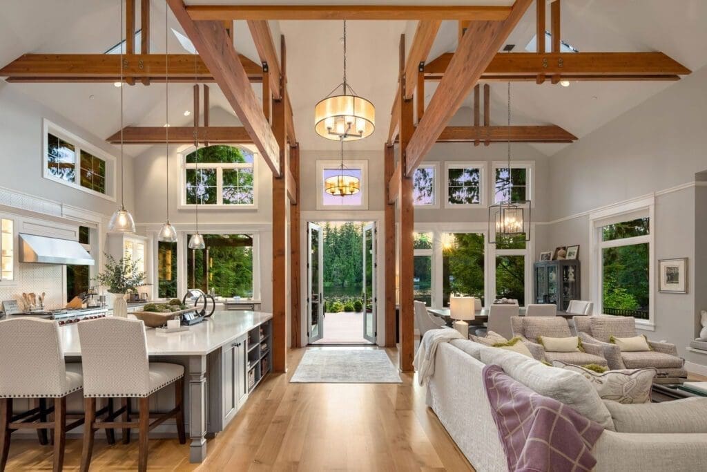 A spacious modern living room with large windows, wooden beams, and high ceilings. Features a kitchen with an island on the left, a cozy seating area with a gray sectional sofa on the right, and a dining area in the center, leading to an open outdoor space.