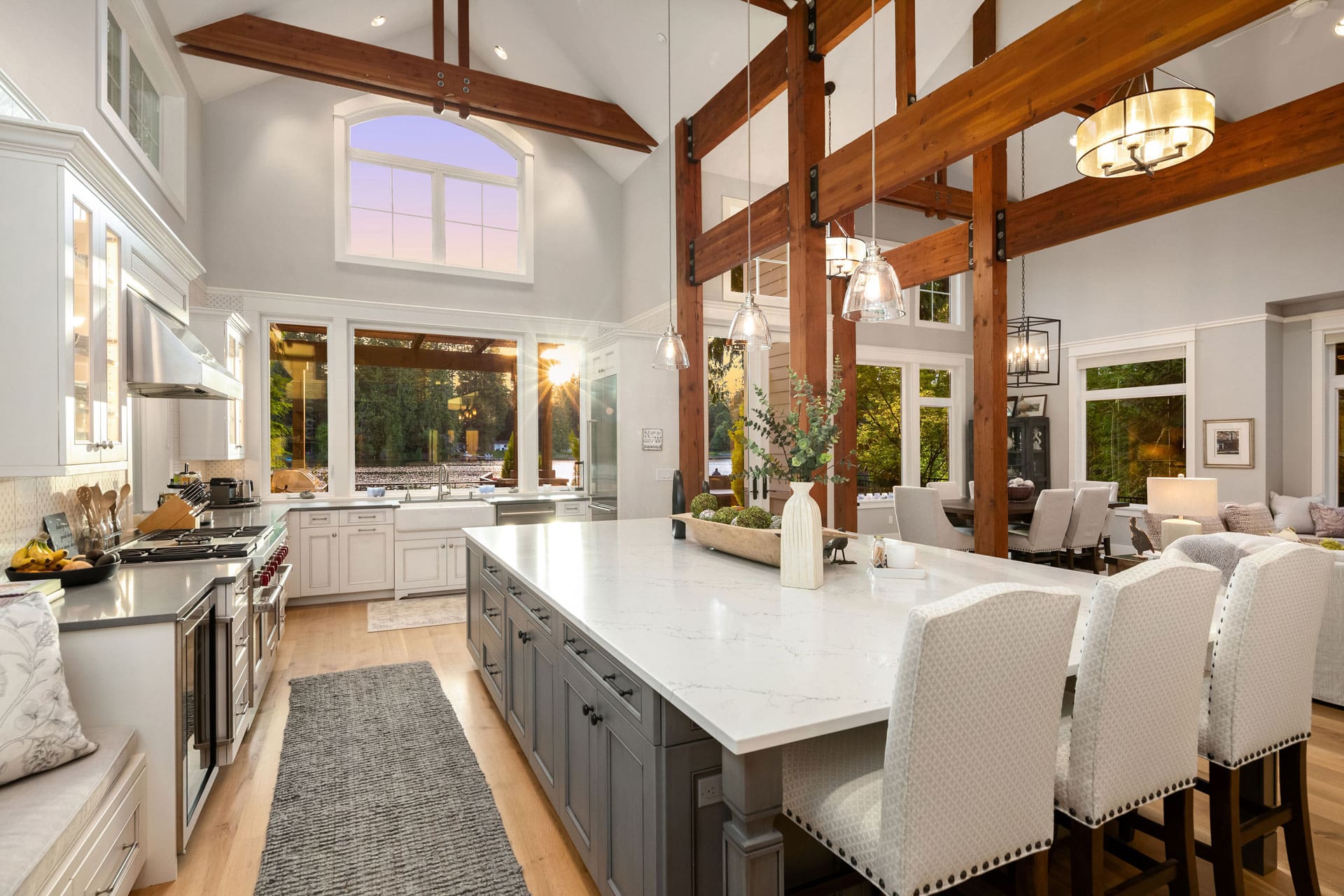 A modern, spacious kitchen and dining area with high vaulted ceilings and large wooden beams. The kitchen features a large island with white countertops, white cabinetry, and stainless steel appliances. Large windows provide ample natural light and a scenic view.