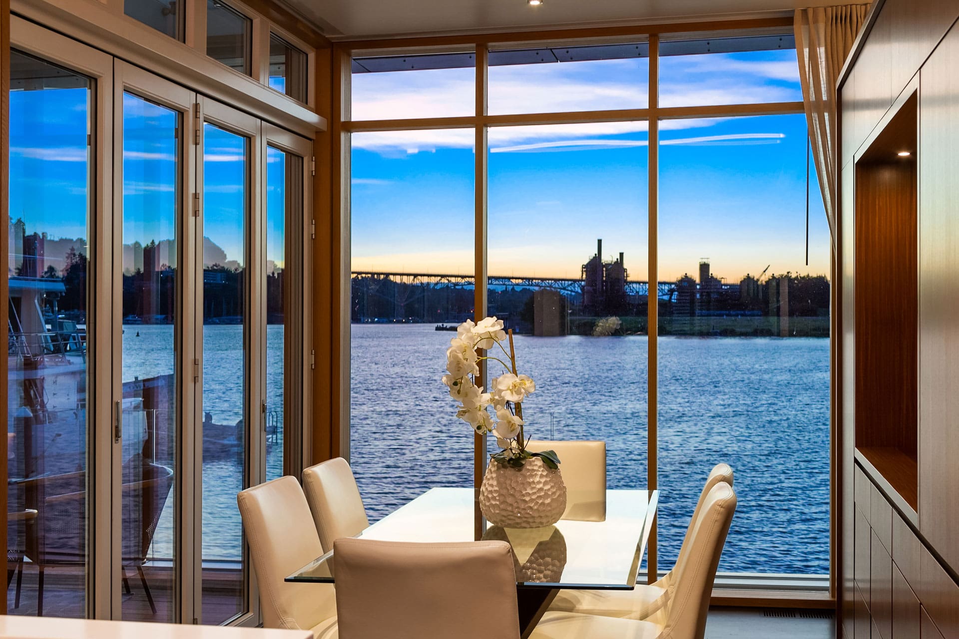 A modern dining room with glass walls overlooks a serene lake at sunset. The table, set with white chairs, is adorned with a white vase holding white flowers. The wall to the right features a large wooden cabinet, adding a warm touch to the sleek design.