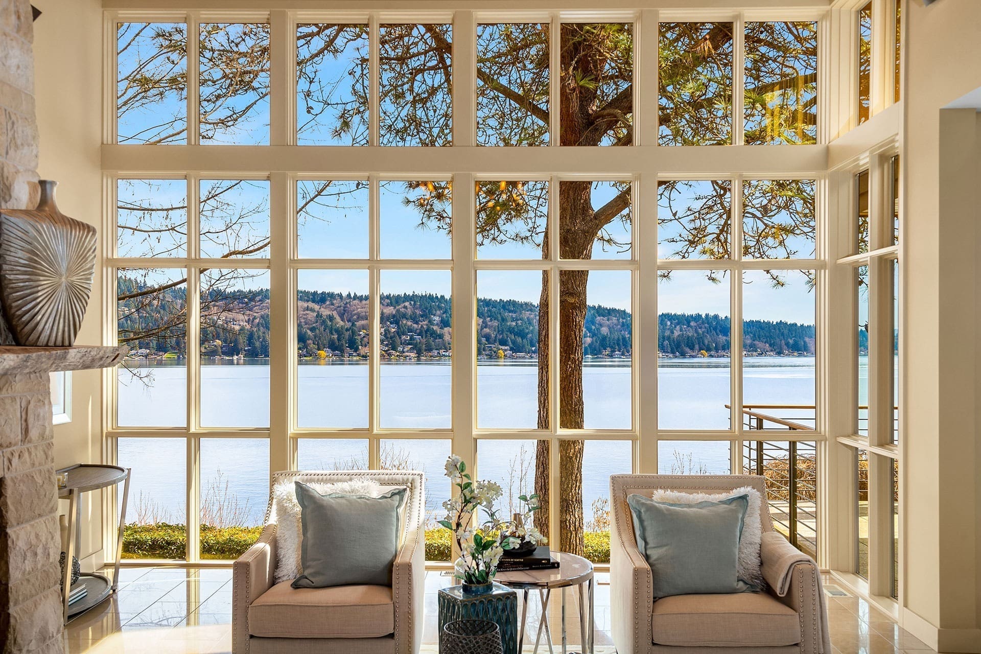 A room with two beige armchairs and a small table between them, positioned in front of large windows overlooking a scenic lake and tree-covered hills. The bright, sunny view provides an open and serene atmosphere, with light streaming through the windows.