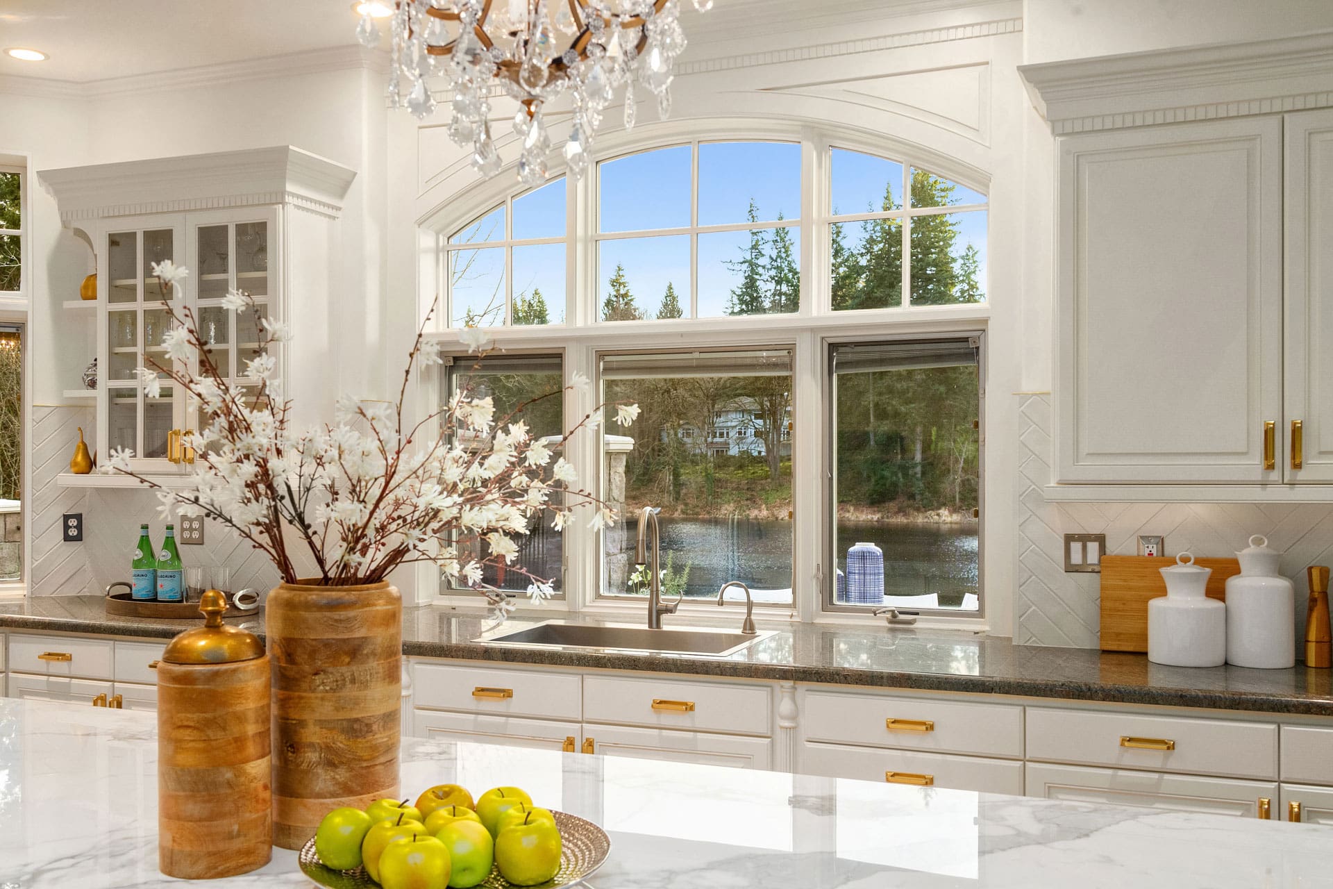 A bright kitchen with large arched windows above the sink, offering a scenic view of trees and a lake. The kitchen features white cabinets, a marble island with a vase of white flowers, wooden canisters, a bowl of green apples, and a crystal chandelier.