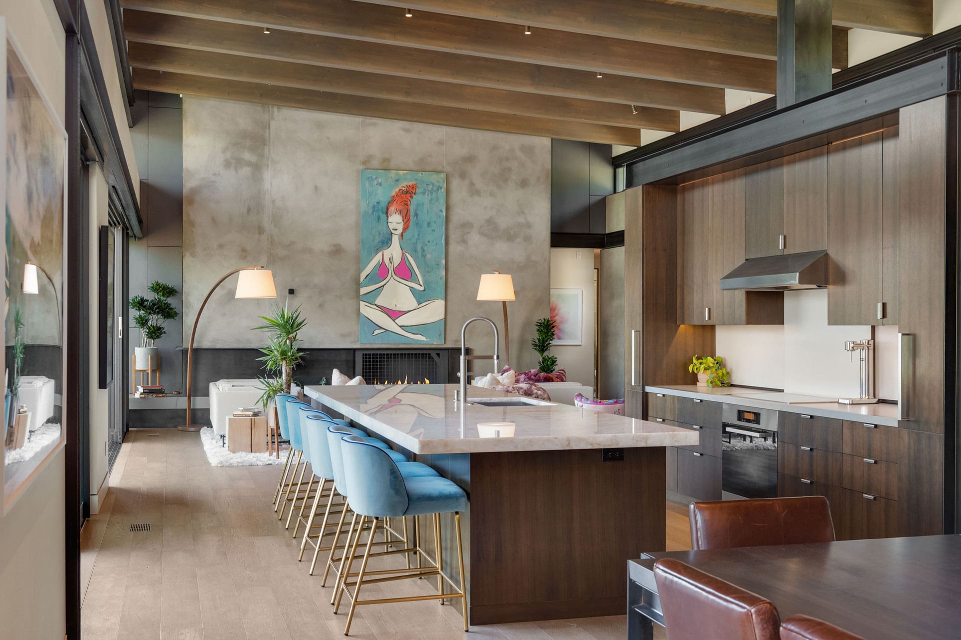 A modern, spacious kitchen and dining area with high ceilings and wooden beams. The kitchen island has blue barstools, and sleek dark cabinetry with white countertops. A large artwork of a figure hangs on the wall, and there's a cozy seating area with a floor lamp.